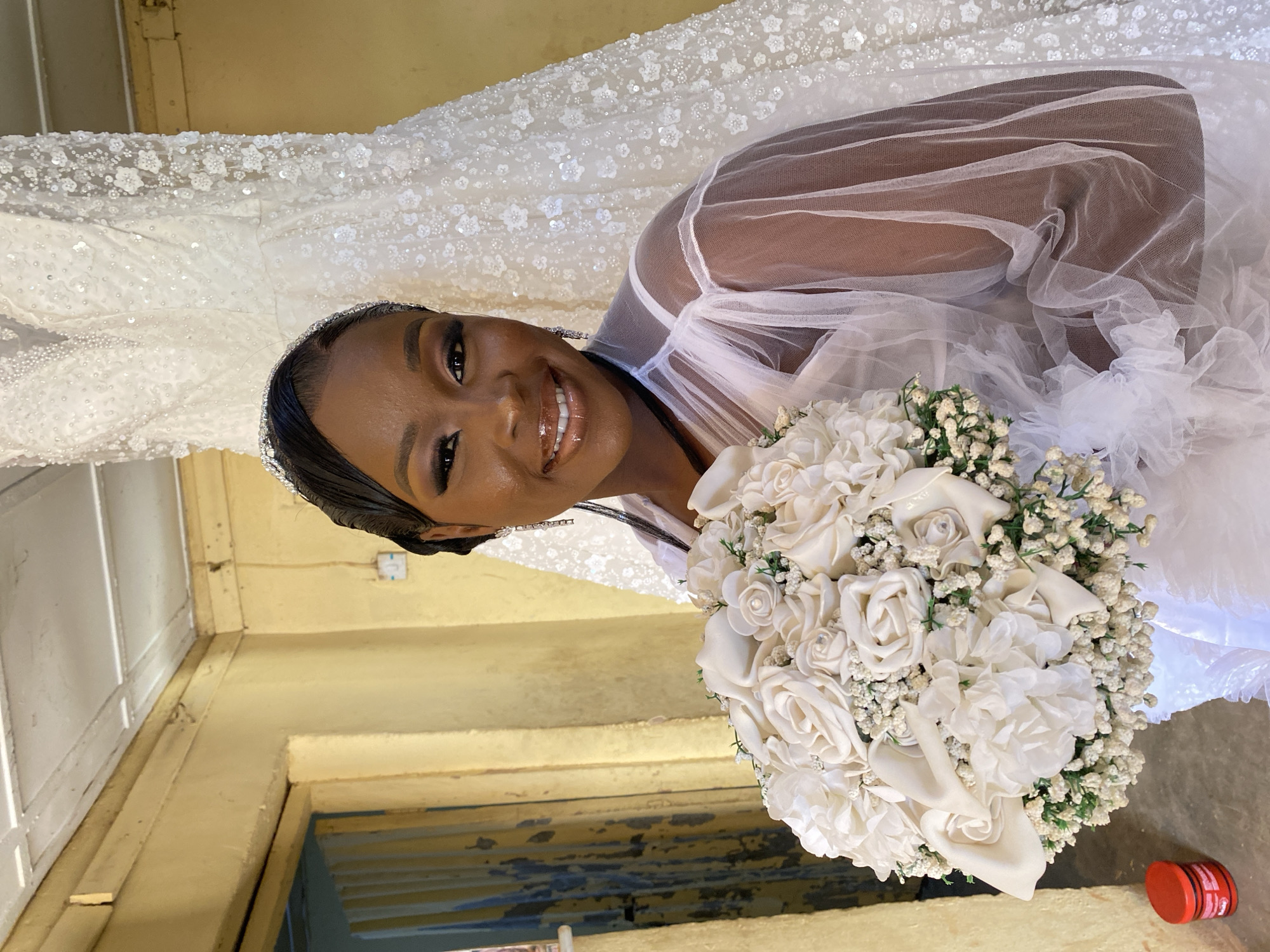 A woman is posing indoors with a bouquet of white flowers. She is wearing a white robe or dress and a sequined headband. The background includes a heavily ornamented white gown or curtain. The interior appears to be a simple room with peeling paint and faded yellow walls. Based on the location Vom, Nigeria provided, the scene likely captures a pre-wedding or bridal photoshoot in a domestic or studio setting.