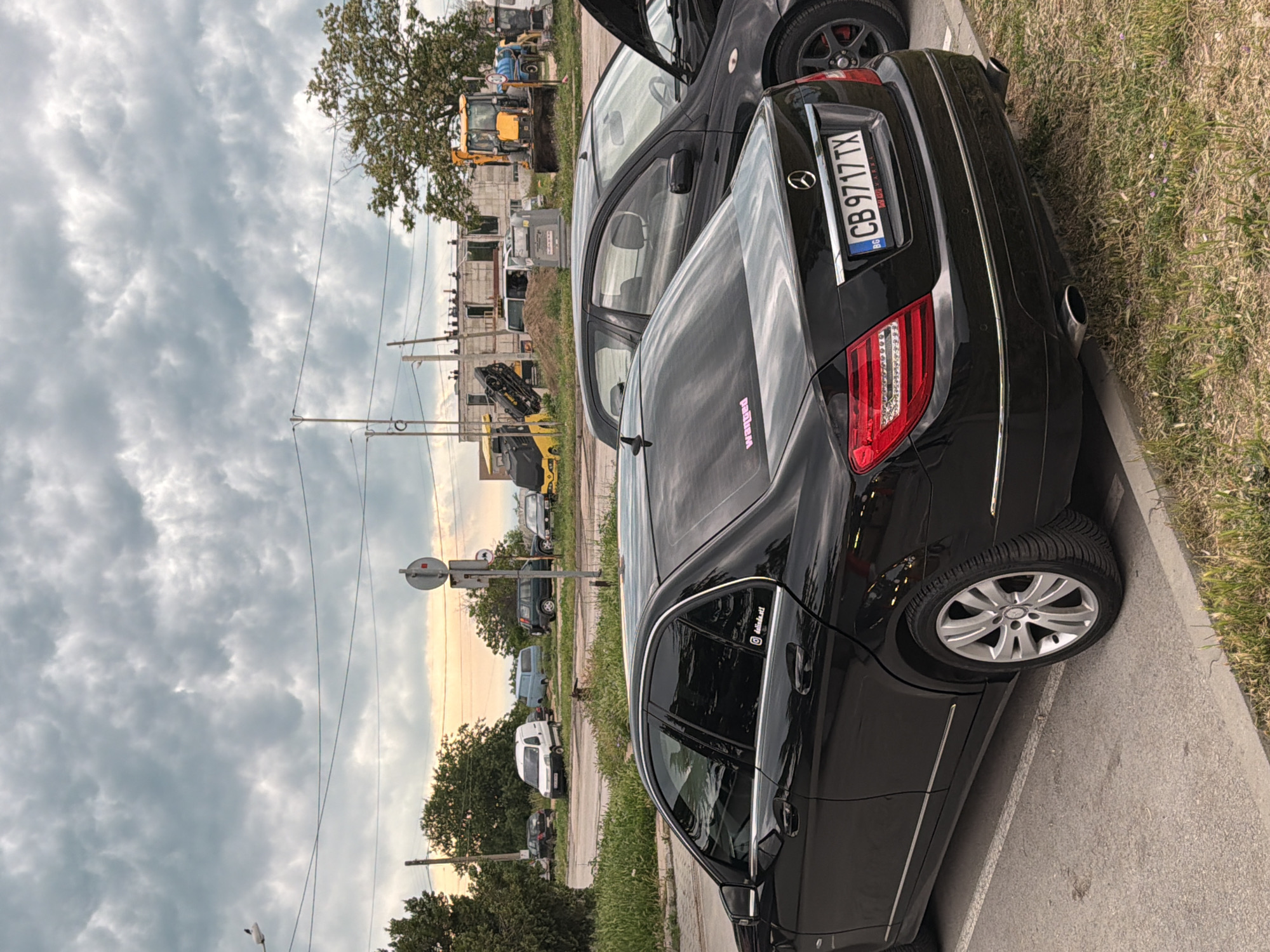 The image, oriented on its side, depicts a street scene under a cloudy sky. In the foreground, a black Mercedes-Benz sedan is parked on the side of a paved road, adjacent to a grassy verge and concrete curb. Its rear end is prominently visible, showing two exhaust pipes and silver alloy wheels. Another black car with an open hood is partially visible in front of the Mercedes.
Utility poles and power lines extend across the mid-ground. In the background, there's an area with apparent construction or industrial activity, featuring several yellow heavy machinery vehicles (excavators/backhoes) and unfinished concrete structures. A large tree with green foliage stands near this site. Further down the road, several other vehicles, including white and blue vans and dark sedans, are parked. A circular street sign is mounted on a pole to the right.
The sky is largely covered with thick, grey clouds, but a strip of warm, lighter sky is visible towards the right horizon, suggesting either late afternoon or early morning.
Visible text includes the Bulgarian license plate "BG CB 9717 TX" on the Mercedes. A pink sticker on the car's rear window reads "wanted," and another sticker on the driver's side rear window displays "@dunkel.t," likely a social media handle. The location context provided is Varna, Bulgaria.