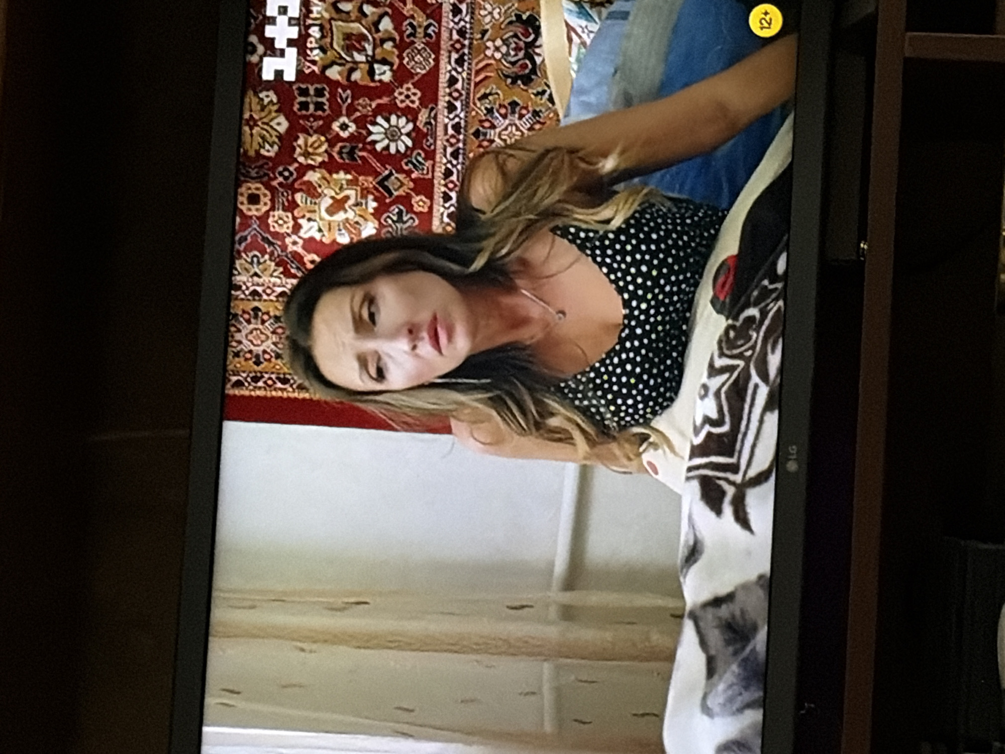 The image displays a medium shot of a woman with long, wavy blonde hair, wearing a black and white polka dot top and a silver pendant necklace. She is seated in what appears to be a bedroom, with a patterned tapestry hanging on the wall behind her. A portion of a blanket with a gray and white pattern is visible in the foreground. The woman's expression is somber, and she is looking off to the side. A "12+" rating icon is present on the right side of the image, suggesting this content is a television broadcast. The scene is lit by artificial light, creating some glare on the screen. The overall setting suggests an indoor environment within a residence.