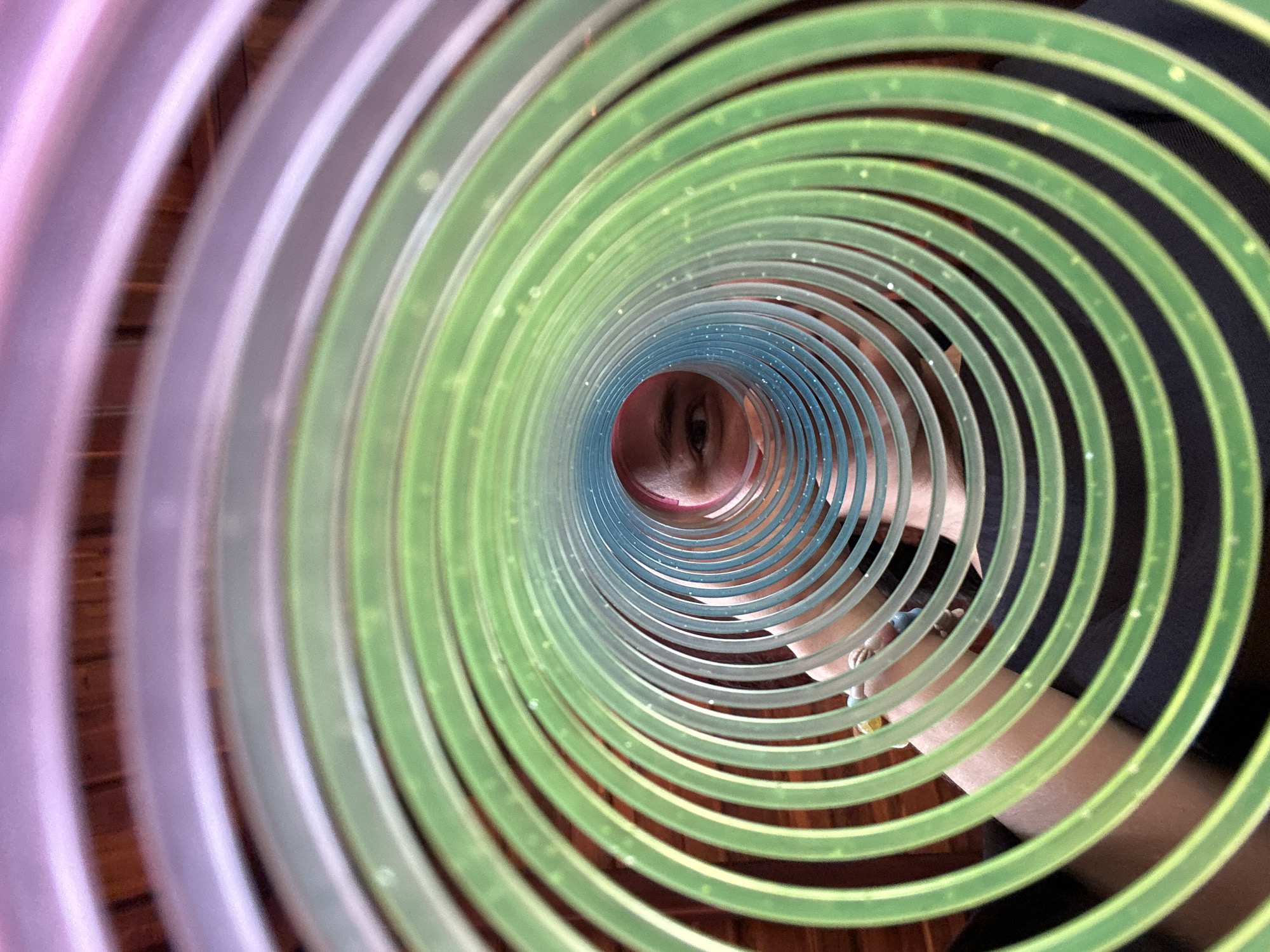 The image depicts a close-up, low-angle view looking through a colorful, spiral-shaped plastic toy or structure. The spiral is composed of concentric rings, transitioning in color from pink and lavender on the periphery to green, then to blue toward the center. At the center of the spiral, a person's face is visible, with a visible eye and part of their mouth. Beyond the spiral and person, there are glimpses of a dark background. No information within the image confirms the location of the shot to be Mũi Né, Vietnam.