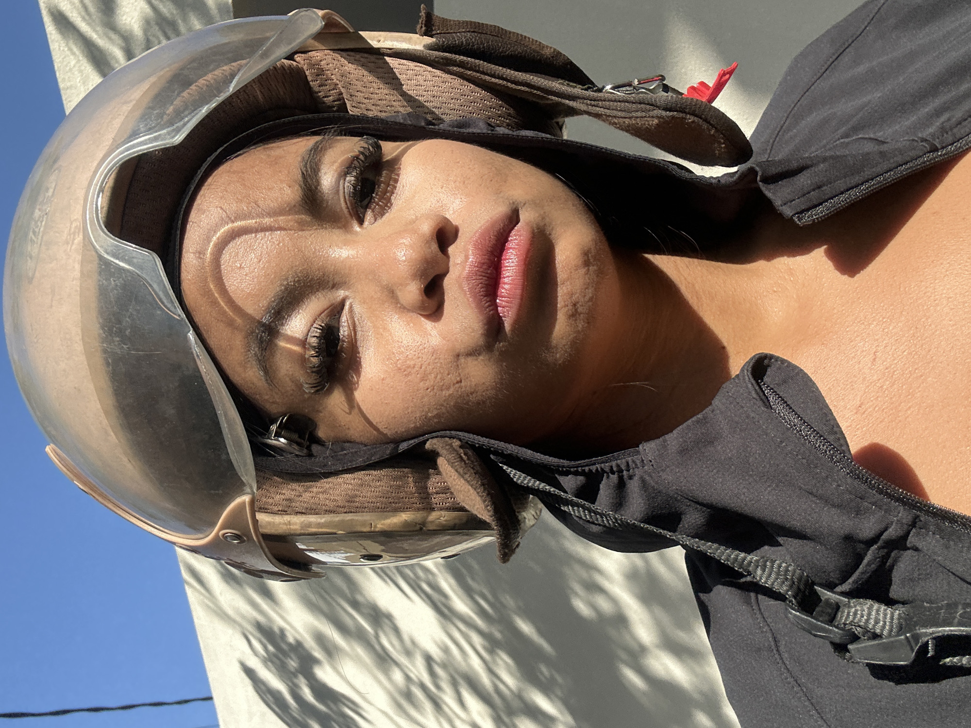 A close-up, waist-up shot shows a young Black woman wearing a brown and tan motorcycle helmet and a black zip-up jacket. She is looking directly at the camera with a neutral expression. Her eyes are adorned with long false eyelashes, and her lips are slightly parted, appearing to have a subtle lip gloss. The helmet has a clear visor that is lifted, and the interior lining is visible. The woman's skin is illuminated by direct sunlight, creating strong shadows on her face and neck. The background consists of a textured, light-colored wall with dappled shadows of foliage cast across it, suggesting an outdoor setting. The sky is visible in the upper left corner, a clear, bright blue. The scene is situated outdoors in Denpasar, Indonesia, on a sunny day.