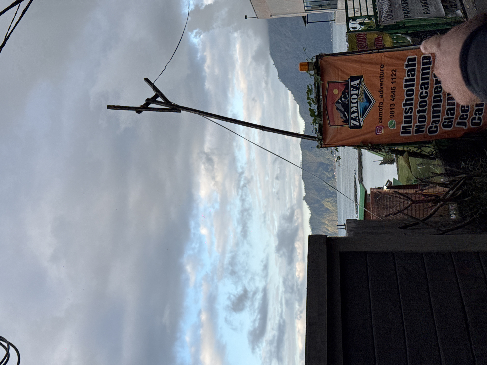 The image depicts an overcast day in Kintamani, Indonesia, with a view of Lake Batur and surrounding hills under a dramatic sky filled with clouds. In the foreground, a dark, textured wall is visible on the left. To the right of the wall, a weathered wooden pole supports electrical wires. A prominent orange banner, advertising "ZAMOFA Adventure," is attached to the pole. The banner features a logo with mountains and water, an Instagram handle "zamofa_adventure," a phone number "0813 4646 1122," and lists services such as "Mushollah, Notocamp, Camper, Glam, Jee, Cai." A person's index finger is visible pointing towards the banner from the bottom right corner of the frame. Beyond the banner, a light-colored building is partially visible on the far right. In the middle ground, several buildings with green roofs are clustered near the water's edge, and a body of water, likely Lake Batur, stretches towards the mountainous horizon. The overall scene suggests a slightly humid and cool atmosphere typical of the Kintamani highlands.