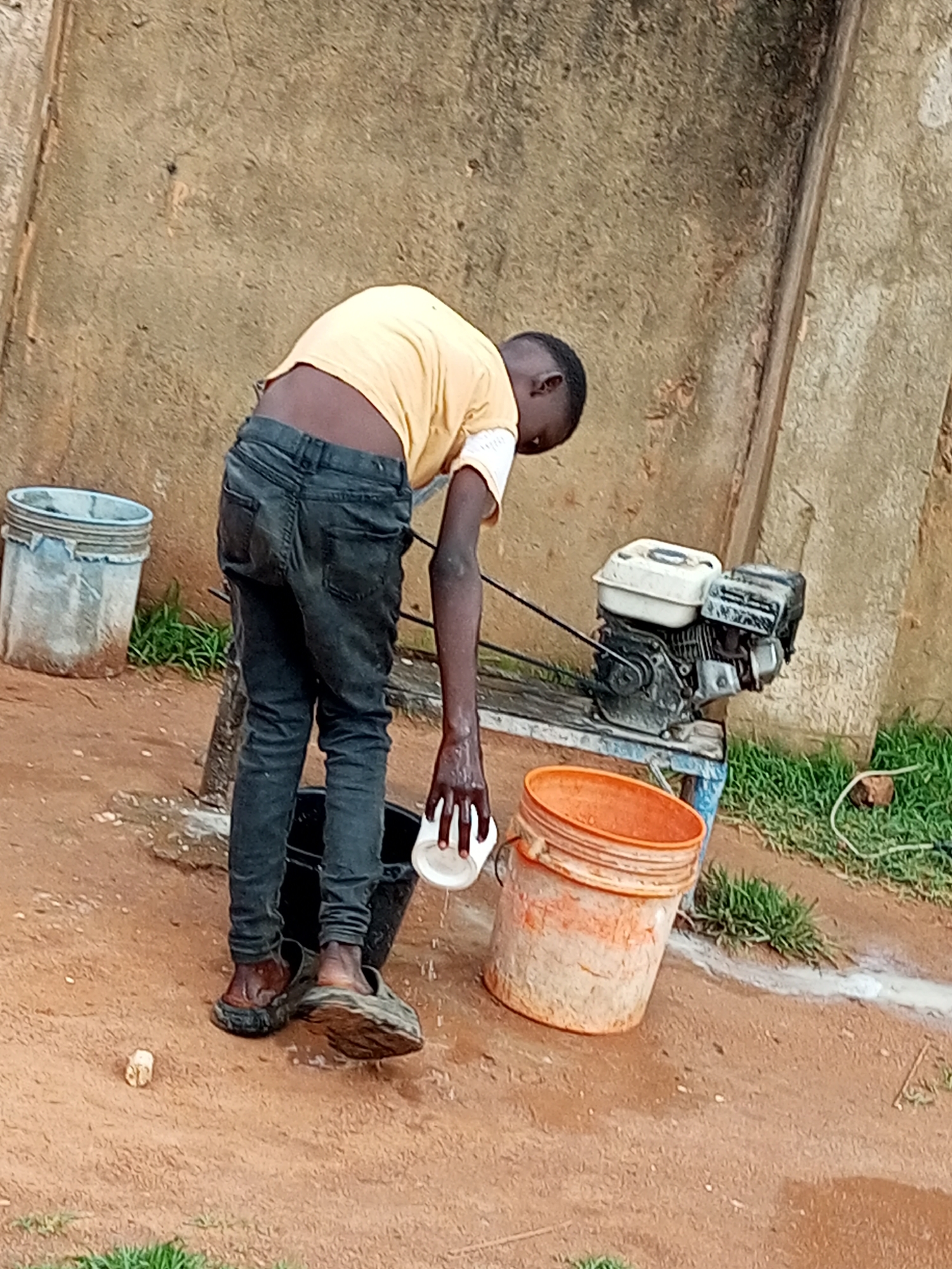 A young person wearing a yellow t-shirt and dark jeans is bent over, pouring water from a white cup into a black bucket. They are standing on a dirt ground, wearing dark clogs on their feet. To the left, a light blue bucket with a grey rim sits on the ground. To the right, a small engine, possibly a generator, rests on a blue and black platform. An orange bucket is also visible to the right of the engine. The background features a textured, light brown wall. The scene appears to be an outdoor area with some grass and a wire. The location is identified as Tempure, Nigeria.