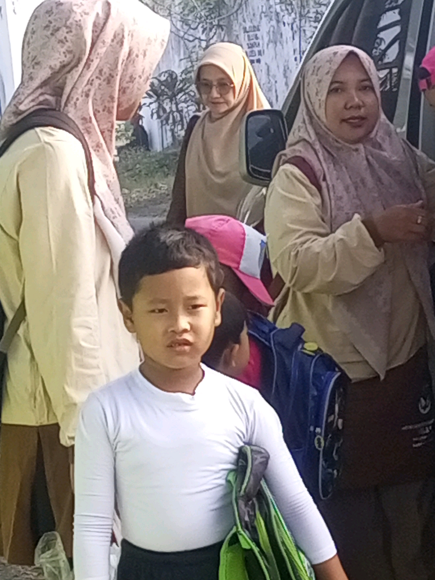In Gurah, Indonesia, a group of people, including a young boy and several women wearing hijabs, stand near a vehicle. The boy, in the foreground, is wearing a white long-sleeved shirt and dark pants, carrying a green and black bag. The women are dressed in modest clothing, with two of them wearing light-colored hijabs and one in a patterned hijab. One woman is partially obscured by the vehicle's side mirror. The environment appears to be outdoors, with a textured wall and some foliage visible in the background. The lighting suggests it is daytime.