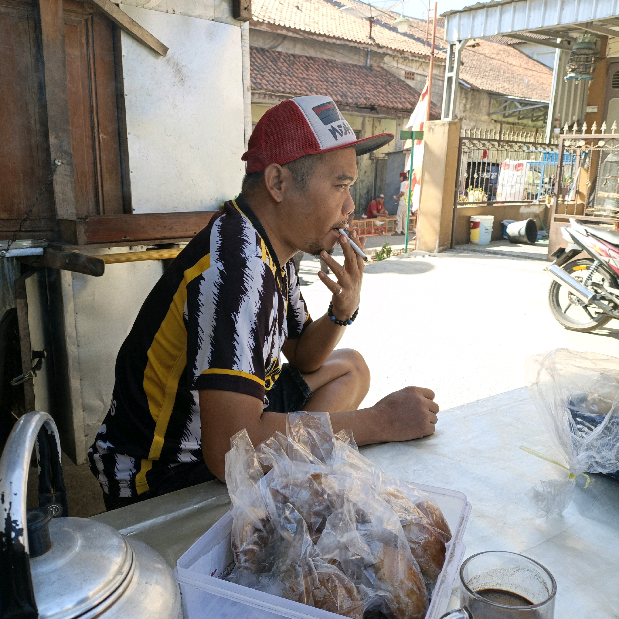 A man in a red and white baseball cap and a black, yellow, and white striped shirt is sitting outside, smoking a cigarette. He is facing right, with his left leg bent and his right leg tucked underneath. In front of him is a table with a glass of coffee, a plastic container filled with bread wrapped in plastic, and a metal kettle on the left side of the frame. In the background, there is a building with a tiled roof, a fence, and a motorcycle. The scene appears to be in a residential area, likely in Randudongkal, Indonesia, given the architecture and general setting. There are other people visible in the background, some standing near a building and others sitting. The lighting suggests it is daytime.