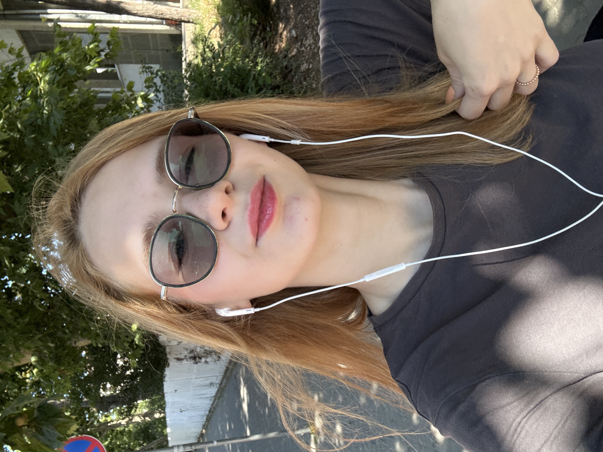 The image shows a close-up, selfie-style photograph of a young woman with long, light brown hair and fair skin. She is wearing round, metal-framed sunglasses with tinted lenses, white earbuds, and a dark gray t-shirt. Her lips are lightly colored with pink lipstick. The background consists of green trees, a white wall or fence, and a paved walkway. The lighting suggests it is daytime, with sunlight casting shadows. A traffic sign with red and blue circles is partially visible on the left. The setting appears to be an urban outdoor environment, likely a sidewalk or park in Kragujevac, Serbia.