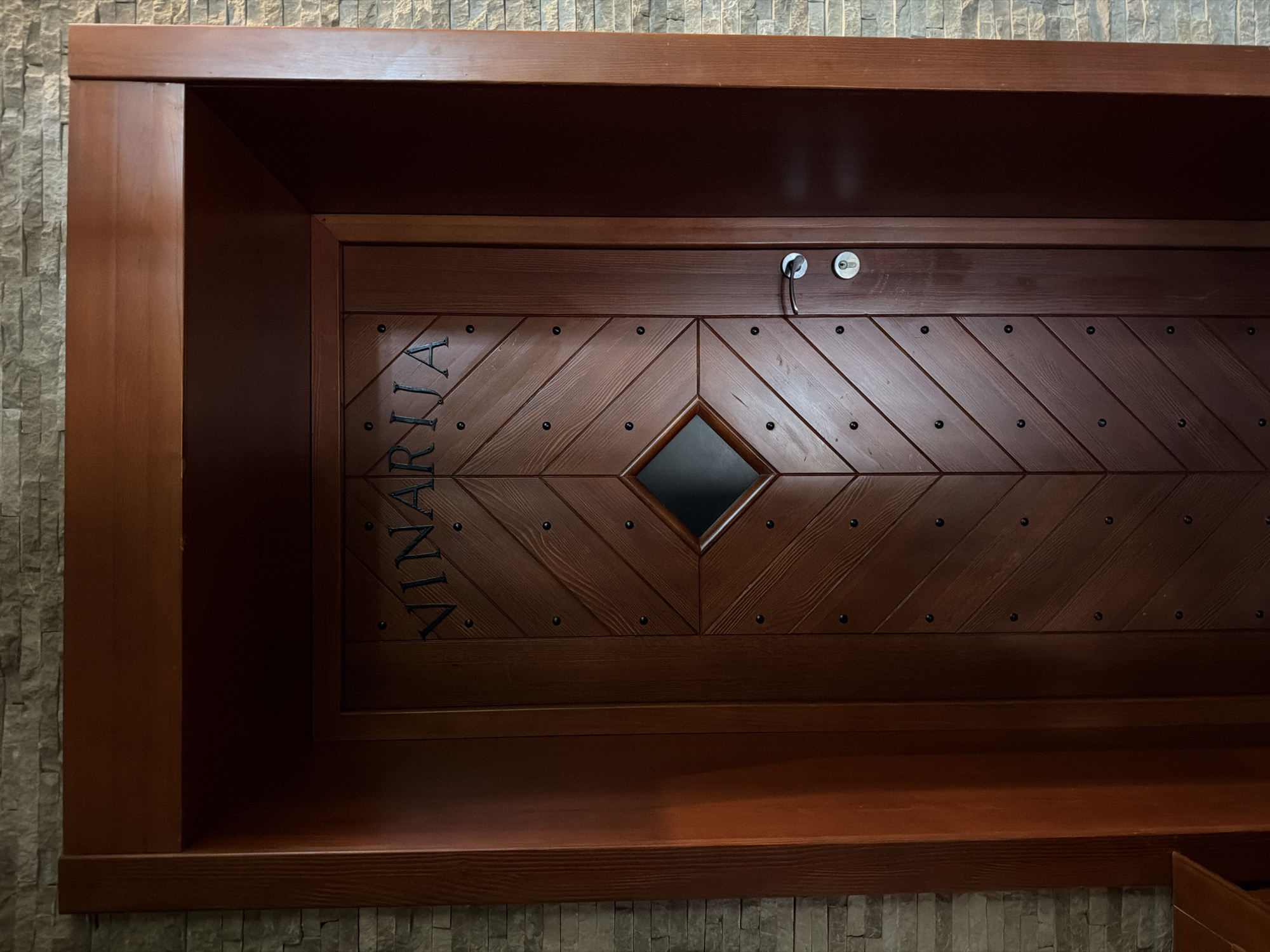 The image shows a dark wooden cabinet in an interior setting. The cabinet's door features a diamond-shaped window in the center and is decorated with parallel grooves and small, round dark metal accents. The word "VINARIJA" is vertically inscribed on the left side of the door. A key is inserted into one of the locks on the top center of the cabinet. The cabinet is positioned against a wall covered with light gray stone-like tiles. Based on the word "VINARIJA", which translates to "Winery," it is highly probable that this object exists at the Vinarija, or Winery, in Krašići, Montenegro.