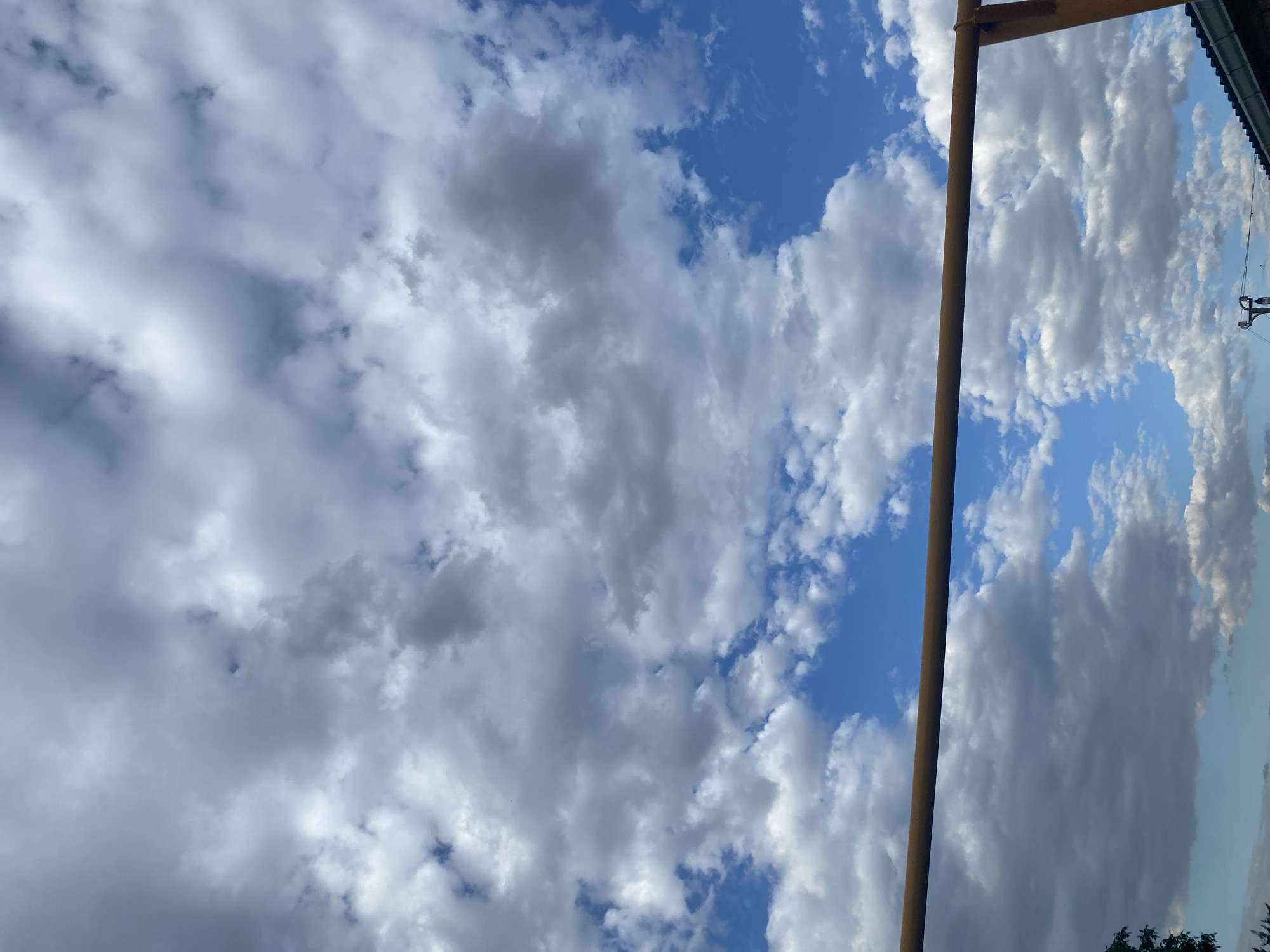 The image captures a bright, overcast sky filled with dramatic, textured cumulus clouds. A thin, rusty metal bar, possibly part of an outdoor structure or play equipment, extends horizontally across the lower portion of the frame, obscuring a sliver of the sky. Below the bar, the edge of a dark, tiled roof is visible on the right side of the image, hinting at a residential or urban setting. A small portion of a green tree appears on the far left. The sky itself is a vibrant blue peeking through the white and grey clouds, suggesting a day with variable weather conditions. While the location is specified as Taganrogsky, Russia, the visual information within the frame does not offer specific landmarks or contextual clues to confirm this geographically.