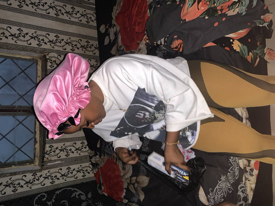 A person lies on a bed with floral patterned sheets. They wear a pink satin bonnet, sunglasses, a white T-shirt with a graphic print, and striped tan and black leggings. The person holds a black package with a white and blue box on top. A window with a patterned wall is visible in the background. There is also a red garment draped on the bed.