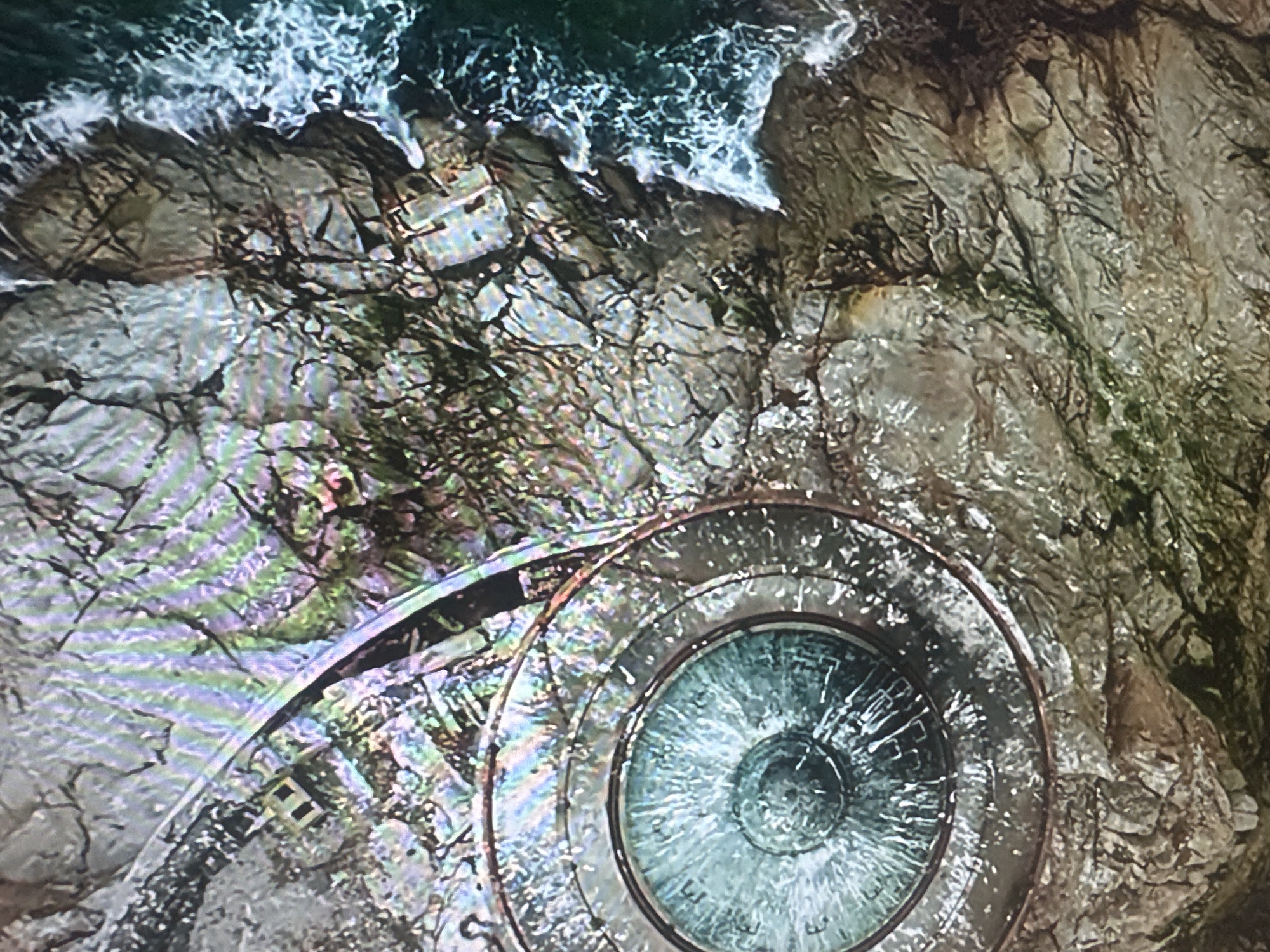 The image is an aerial view of a circular, glass-covered observation deck situated on a rocky coastline, with waves crashing against the shore. The observation deck has a metallic ring around its perimeter and appears to offer a view of the water below. The surrounding landscape is rugged and rocky, with hints of vegetation or moss growing on the rocks. There are no people visible in the image. The location is identified as the city of Almaty, Kazakhstan.