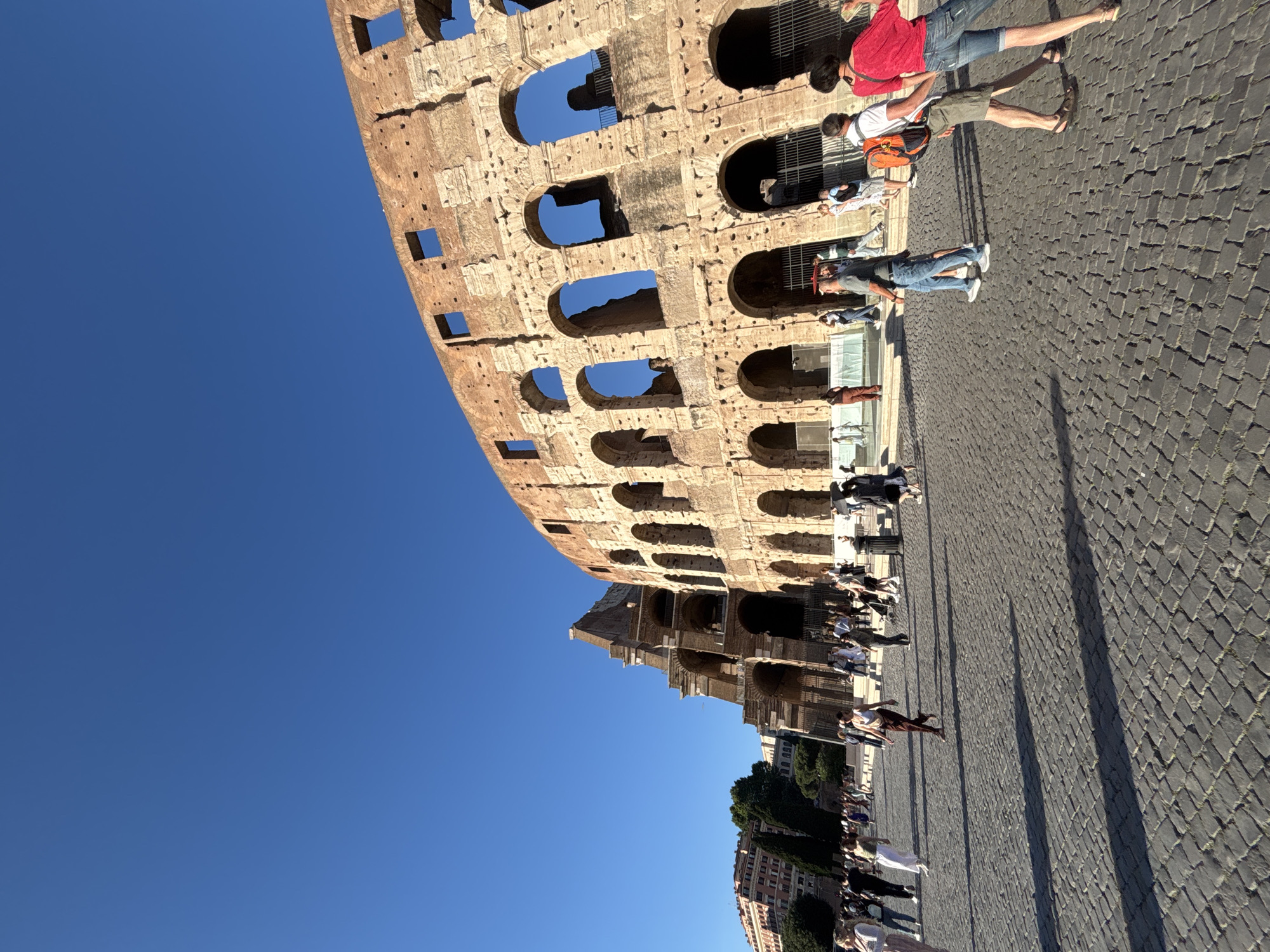 This image shows the Colosseum in Rome, Italy, from an external perspective. The structure is composed of layered arches and openings, partially showcasing the clear blue sky behind it. In front of the Colosseum, several people are walking and standing on the cobblestone ground. Some are close to the building's walls, while others are further away. Adjacent to the Colosseum, some greenery and buildings are visible, indicating the surrounding urban environment.
