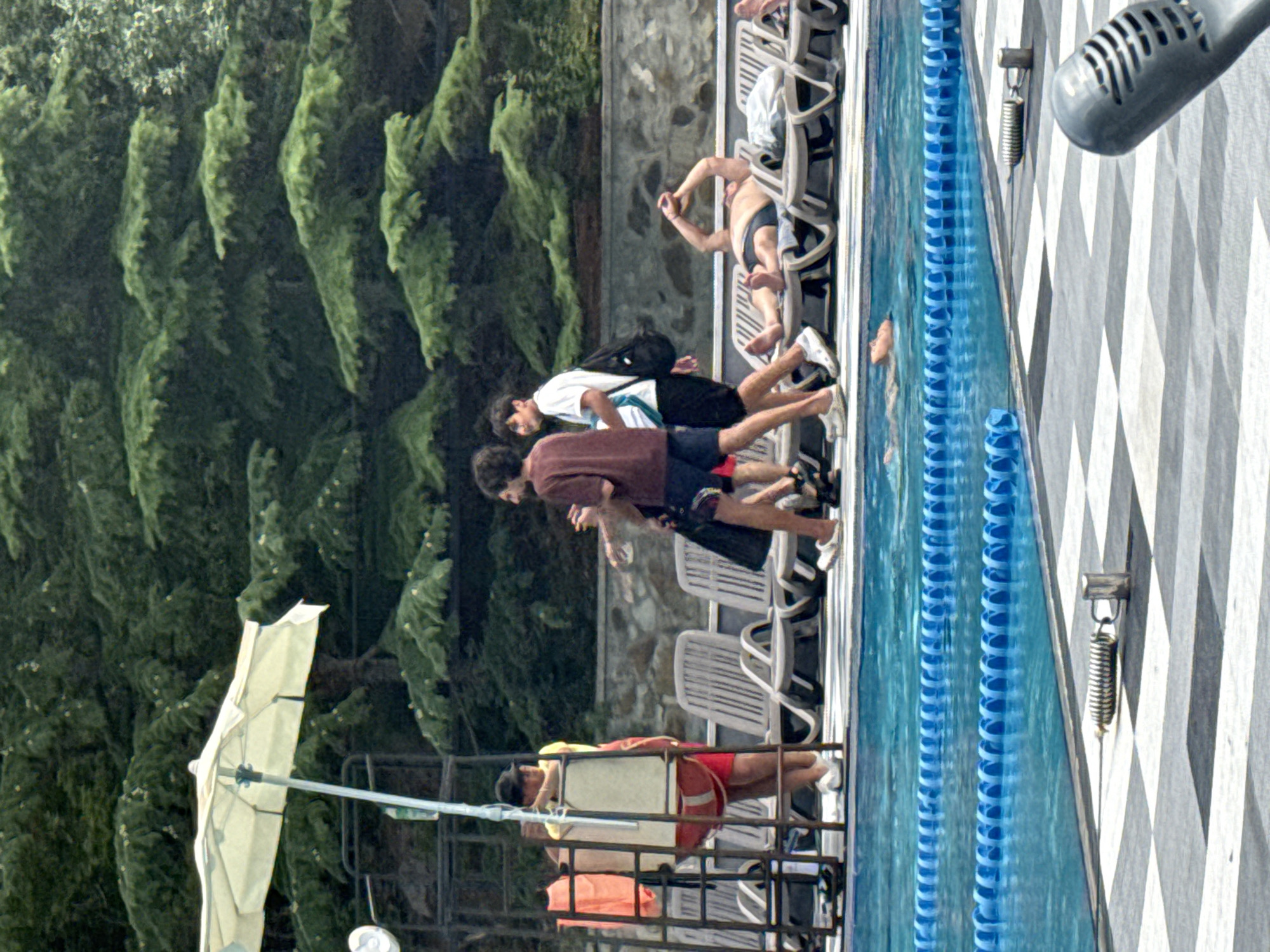 The image depicts a poolside scene with several individuals and objects. In the foreground, a swimming pool with lane markers is visible, with a checkered pattern on the pool deck. A detached microphone is positioned on the deck in the lower right corner. In the middle ground, three men are walking near a lifeguard stand topped with a partially unfurled white umbrella. One man in the foreground wears a maroon shirt and dark shorts, another in a white t-shirt and dark shorts carries a black backpack, and a third man is partially visible in the background. Beyond the lifeguard stand and the walking men, several lounge chairs are arranged. One person is lying on a lounge chair with their arms raised. In the background, lush green pine trees form a dense forest. A stone wall is visible behind the lounge chairs. The scene appears to be an outdoor recreational area, possibly a hotel or resort, in a location with a dense forest environment. The location is identified as Массандровский поселковый совет, Ukraine.