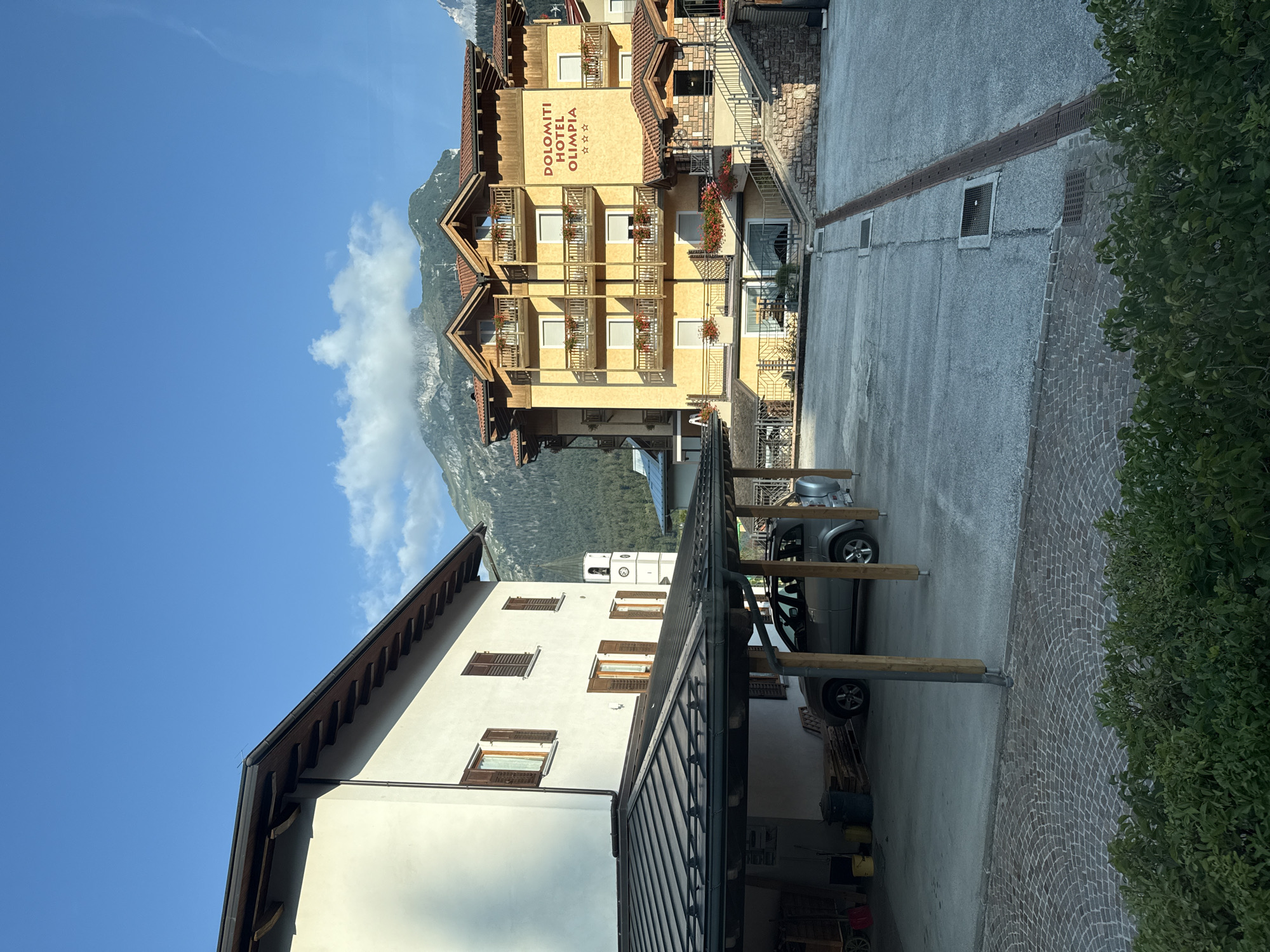 The image depicts a street scene in the city of Andalo, Italy, under a clear blue sky with scattered clouds. In the foreground, lush green foliage lines the bottom of the frame. The midground features a paved area with a drainage grate and a patterned brick walkway. To the left, a white building with brown shutters is partially visible, with a covered parking area in front of it sheltering a silver SUV and some wooden pallets. A white clock tower with a green roof stands behind this building. Dominating the right side of the midground is a yellow hotel building with a red roof, adorned with balconies and flower boxes. The hotel's signage reads "DOLOMITI HOTEL OLIMPIA" with three stars below it. A stone staircase leads up to the hotel's entrance. In the background, a majestic mountain range with some trees and snow-capped peaks rises against the sky. The overall scene suggests a tranquil and picturesque mountain resort town.