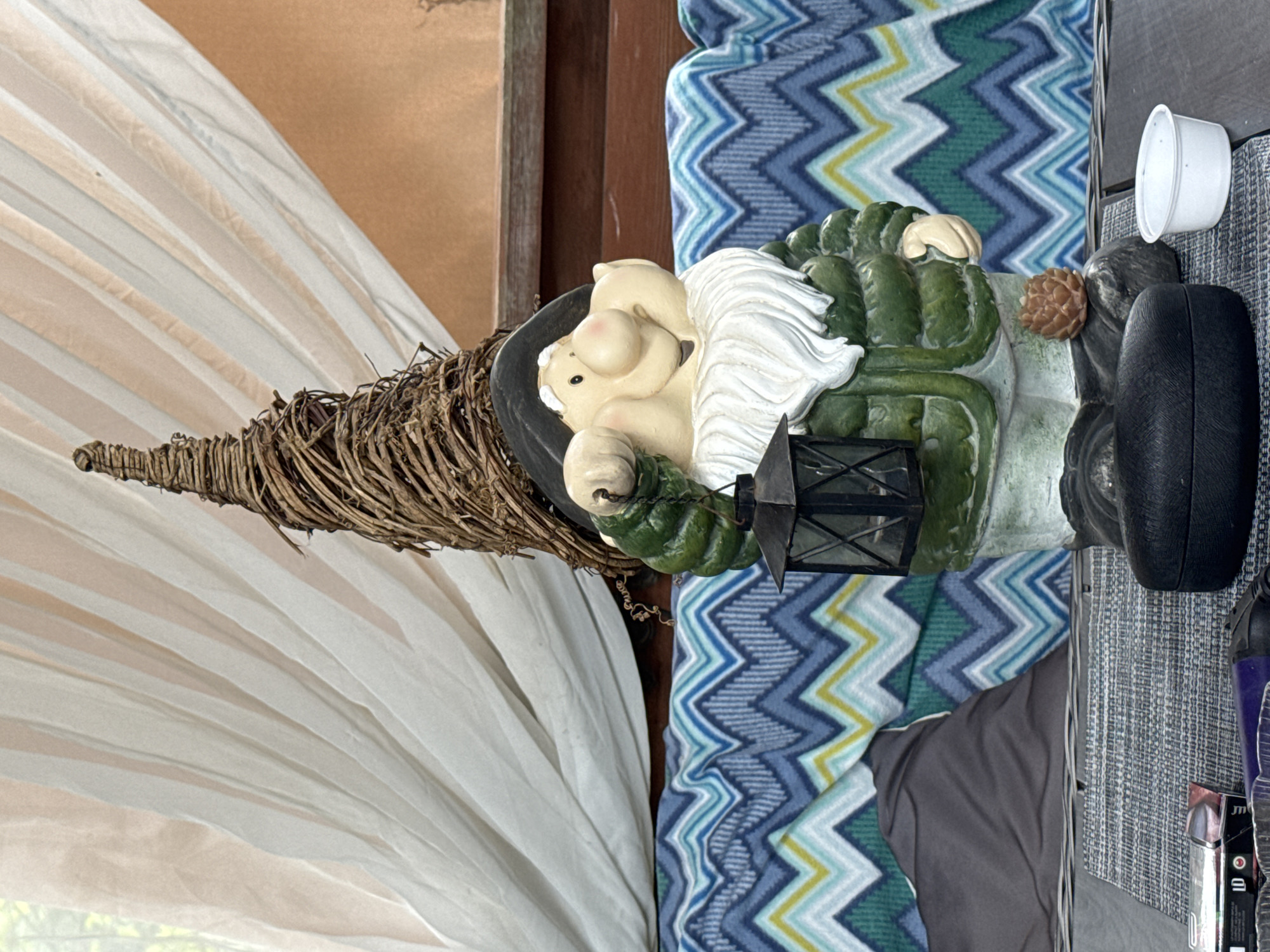 A gnome garden statue stands in front of a blue and white chevron pattern blanket. The gnome is wearing a green jacket, a black hat, and has a white beard. It is holding a black lantern in its right hand. The gnome's hat is conical and made of twigs. Behind the blanket, there is a white curtain. In the foreground, there is a gray table with several objects on it, including a small white container and what appears to be a dark case. The scene is outdoors, possibly on a patio or deck. The image does not contain any explicit location information for Bersenevka, Russia.