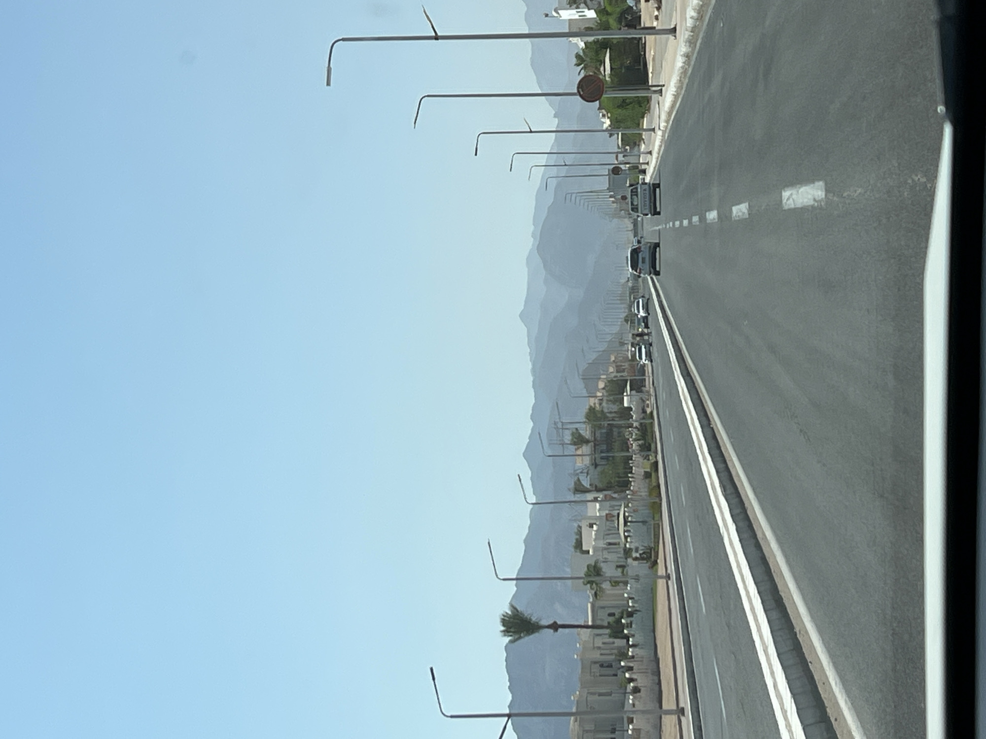 The image depicts a wide, multi-lane asphalt road in Sharm El Sheikh, Egypt, likely a major thoroughfare given its size and the presence of streetlights on both sides. The road is lined with modern, low-rise buildings in a light color, suggesting a residential or commercial development. Palm trees are visible interspersed with the architecture, and a backdrop of hazy, layered mountains stretches across the horizon under a clear, pale blue sky. Several vehicles, including cars and a white pickup truck, are traveling on the road. The overall impression is one of a sunny day in a developing urban or suburban area with a mountainous landscape.