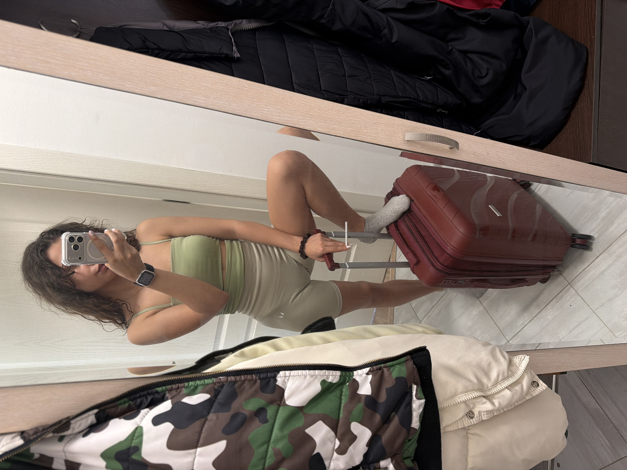 The image is a mirror selfie taken indoors, rotated 90 degrees clockwise. A young woman with long, dark, wavy hair stands in what appears to be a room, possibly a hotel or bedroom, reflected in a large mirror with a wooden frame. She is wearing a light green athletic crop top and matching light-colored athletic shorts or bike shorts. On her left wrist, she wears a black smartwatch, displaying the time as "2:20". She holds a white smartphone with a clear case in her left hand, pointed towards the mirror to capture her reflection. In her right hand, she holds a white object, possibly a cigarette or vape pen.
Her right leg is bent, with her foot, clad in a white patterned sock, resting on a red hardshell suitcase that has its handle extended. The suitcase is positioned on a light-colored tiled floor, also visible in the reflection.
Above the mirror's reflection, in the foreground (or bottom of the original frame), a camouflage patterned puffer jacket and a light beige puffer jacket are visible, lying on a surface. In the background of the reflection (or top of the original frame), a dark, possibly black, quilted jacket is draped over another surface. The overall setting suggests a transient environment, such as a hotel room, perhaps during travel or packing.