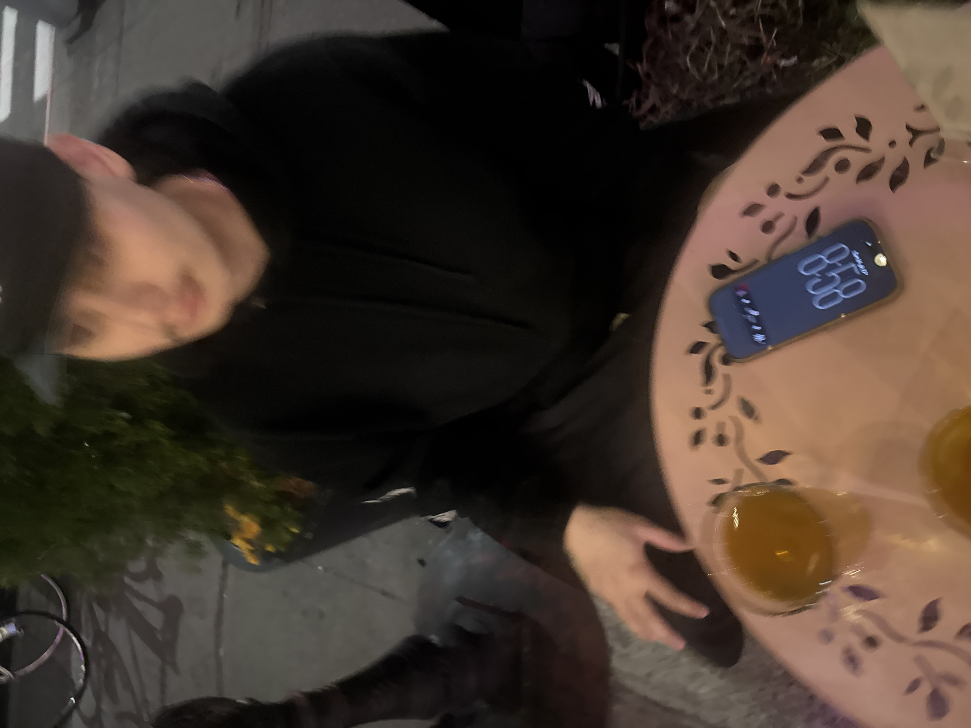 The image captures a person in a black hoodie and baseball cap seated at an outdoor table. The person is slightly out of focus but appears to be fair-skinned. The table, adorned with a floral cut-out design and a pinkish hue, holds a smartphone displaying the time 8:58 and two drinks in glasses. The background shows a sidewalk and greenery, with a hint of what appears to be a bicycle frame. The image does not contain enough details to confirm the location in the city of New York, United States of America.