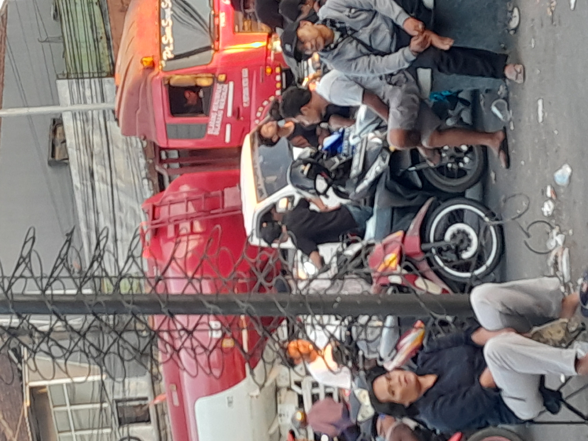 This image depicts a busy street scene in Gentengwetan, Indonesia, likely during daylight hours. In the foreground, a woman is seated on a folding chair, wearing a dark long-sleeved shirt and light-colored pants. Her gaze is directed slightly to her right. Adjacent to her, several motorcycles are parked, some with passengers.
In the mid-ground, a line of motorcycles and their riders is visible, some engaged in conversation. A white van is positioned behind them. Further in the background, a large red tanker truck dominates the scene, with the text "Pt. Sian Soy Sang" visible on its side. Another red truck is also present, partially obscured. The environment appears to be an urban setting with buildings and utility poles visible. The ground is paved and littered with debris.