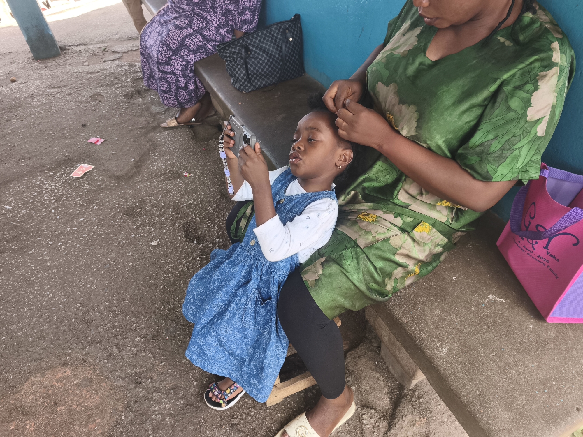 A young girl sits on a woman's lap while her hair is being braided outdoors in Jos, Nigeria. The girl is holding a small electronic device and is dressed in a denim dress over a white long-sleeved shirt, with black leggings and sandals. The woman braiding the girl's hair wears a green floral print dress. They are sitting on a concrete bench next to a blue wall. A purse and a pink tote bag rest on the bench. Another woman wearing a patterned purple dress sits on the same bench. The ground is asphalt.