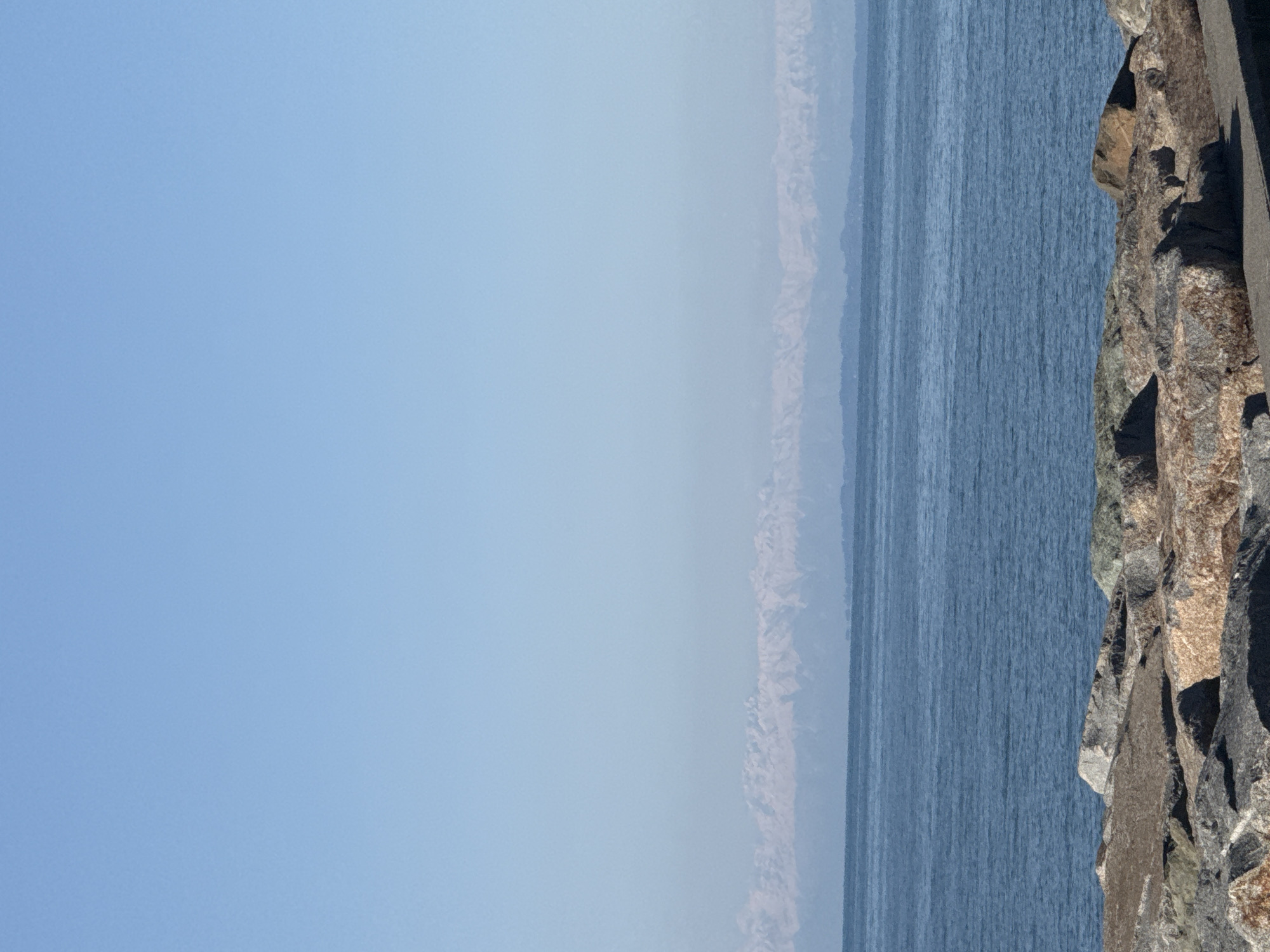 The image presents a serene coastal landscape, offering an expansive view of the sea and sky alongside a rugged shoreline. The upper portion of the frame is dominated by a clear, bright blue sky, indicating fair weather conditions. Below the sky, a vast expanse of calm, deep blue water stretches across the mid-ground, showing subtle textures from gentle ripples or small waves on its surface.
To the far right, a prominent rock formation or cliff face with a textured, jagged surface creates a strong foreground element. These rocks display a variety of natural colors, including shades of brown, grey, and ochre, suggesting a weathered and ancient geological structure. In the distant background, across the water, a faint and hazy light-colored landmass, possibly mountains or a distant coastline, is visible on the horizon, adding depth to the scene.
The photograph captures a peaceful moment in nature. The time of day appears to be daytime, likely mid-morning or afternoon, given the clear skies and lack of harsh shadows. The weather is clear and calm, with no visible clouds or adverse conditions. There are no people, animals, or man-made objects actively engaged in any event or activity. No text is visible within the image. Given the provided context, the setting is Batumi, Georgia, suggesting this is a view of the Black Sea and its surrounding natural environment.