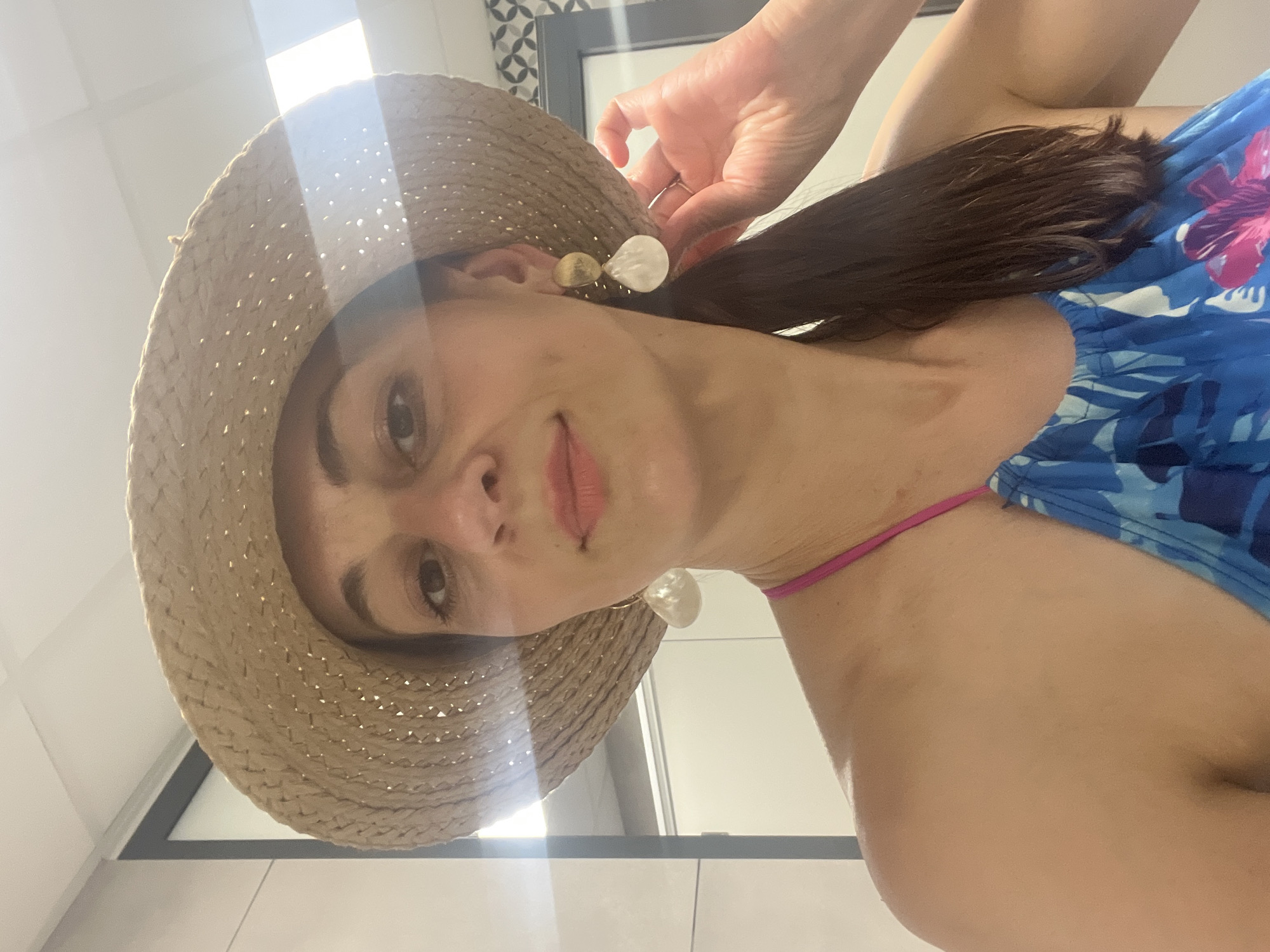A medium shot captures a woman from the chest up, wearing a straw hat that partially obscures her face. She has dark hair pulled back and is wearing a blue and pink floral patterned top. Her right hand is raised, touching her hair near her ear, and she is wearing large, dangling earrings. The background appears to be an indoor setting with tiled walls and bright overhead lighting. The scene is well-lit, with a noticeable glare from the lights on the hat and the woman's skin.