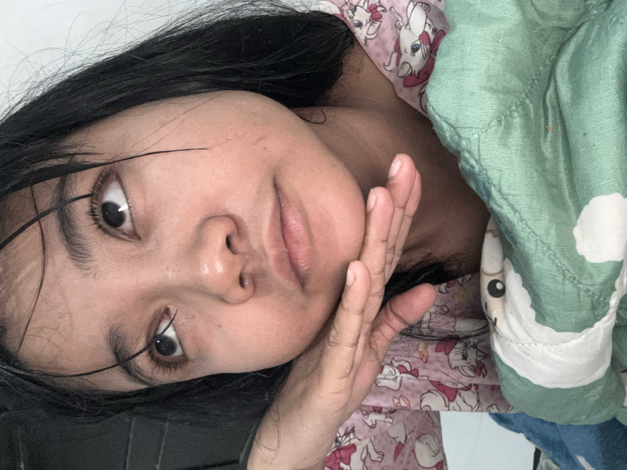 A close-up, indoor, eye-level shot shows a young person resting their chin on their hand. The person has dark hair, which appears wet and is falling across their forehead and face. Their eyes are looking upward, and they have visible eyelashes. They are wearing a pink shirt with a white and pink cat pattern. In the foreground, a green, textured blanket is visible, with a hint of a blue blanket underneath. The background is a plain, white wall. The scene suggests a relaxed or contemplative moment, possibly in a bedroom. The location is identified as North Jakarta, Indonesia.
