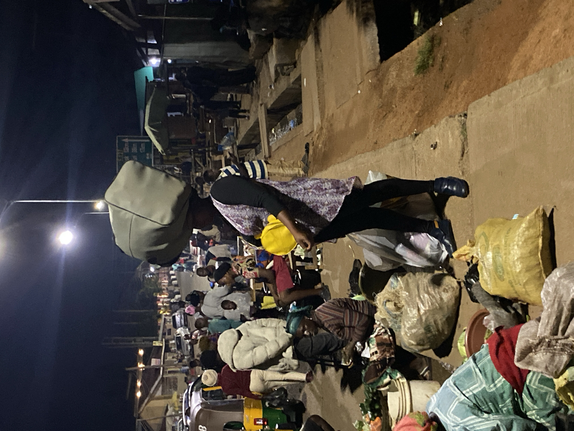 A woman carrying a large, light-colored bag on her head walks along a dirt path next to a group of people and market stalls at night in Bukuru, Nigeria. She wears a patterned dress and black pants, and a yellow bag is held in her right arm. The area appears to be a busy marketplace with various goods displayed on the ground, including produce and sacks of items. Several people are visible, some sitting and others standing, amidst the dimly lit surroundings illuminated by a street lamp. The environment includes a road, buildings, and a drainage ditch.
