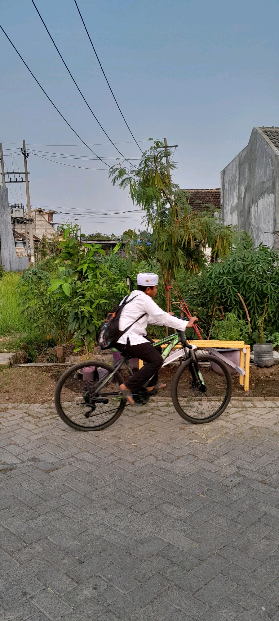 An Indonesian boy wearing a white peci and a white long-sleeved shirt rides a bicycle on a brick pathway. He has a black backpack slung over his left shoulder. The bicycle is dark colored with green accents on the frame. The boy is in motion, as indicated by the slight blur of the wheels. The background is filled with lush green foliage, including trees and bushes. In the distance, utility poles with wires are visible, as well as the side of a concrete building with a tiled roof. The sky is overcast. The scene appears to be in a residential area of Bendungan, Indonesia.