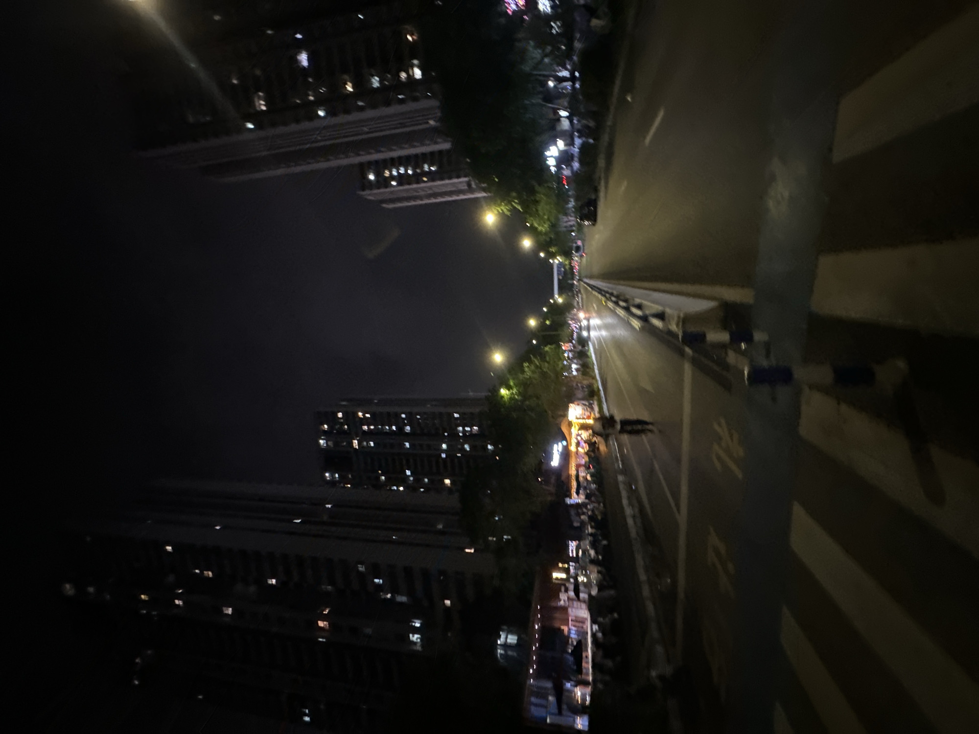 The image captures a nighttime street scene in Chongqing High-tech Zone, China, featuring tall, illuminated apartment buildings on both sides of a multi-lane road. The road surface appears wet, reflecting the streetlights and headlights of distant vehicles, suggesting recent rain. On the left side of the road, a row of establishments with awnings and outdoor seating is visible, some with lights indicating they are open. A lone pedestrian walks across a crosswalk in the foreground, illuminated by streetlights. The sky is dark, with subtle cloud cover obscuring any stars or moon. The overall impression is one of urban nighttime activity, with a blend of residential and commercial spaces.