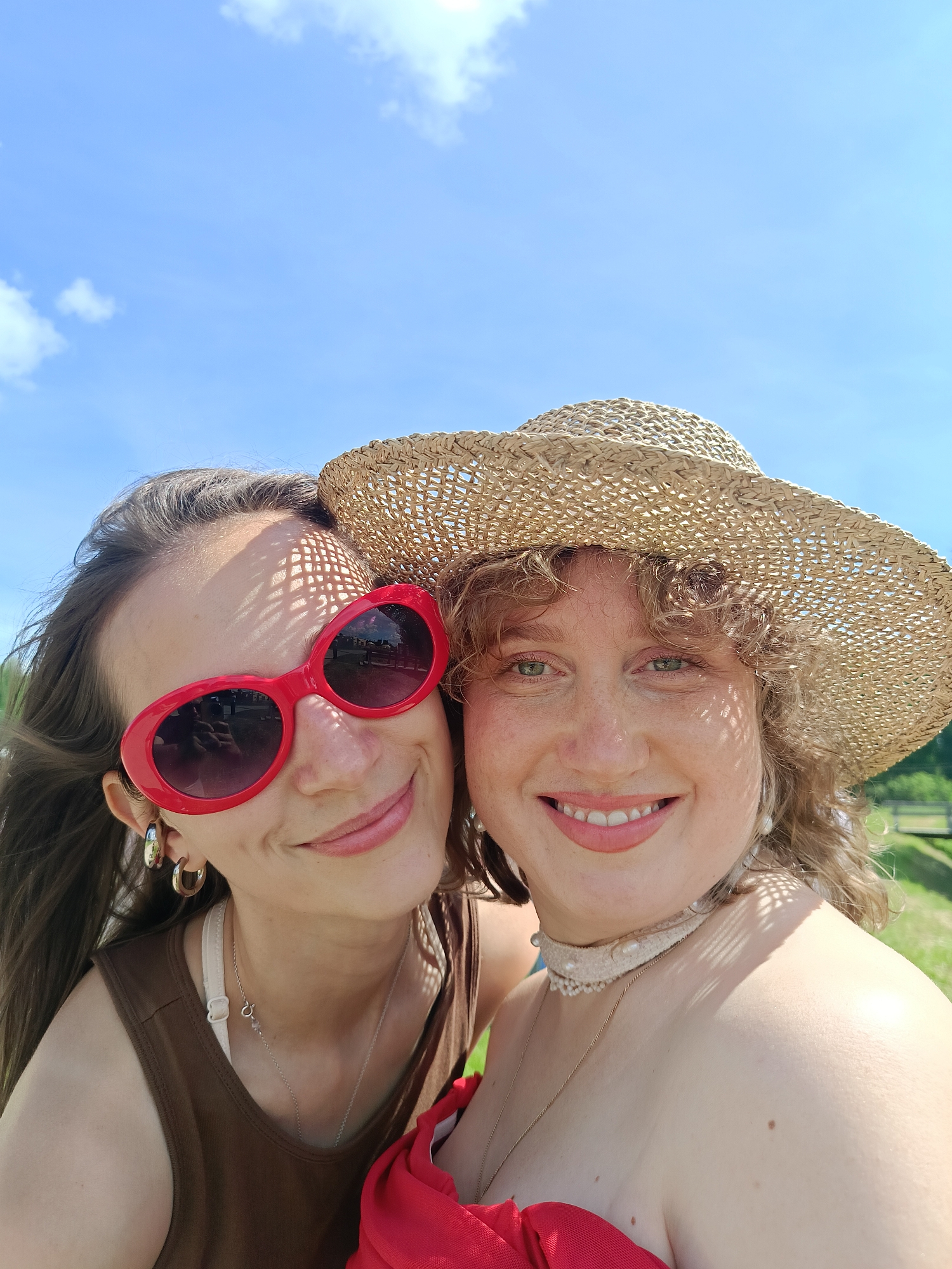 Two individuals are captured in a close-up, selfie-style photograph against a bright blue sky with scattered clouds. The person on the left has long brown hair, is wearing large red sunglasses, and a brown tank top with a white bra strap visible. They are also wearing large hoop earrings. The person on the right is wearing a straw hat and a white lace choker with pearls. They have curly blonde hair and a light complexion with freckles. The background suggests an outdoor, possibly rural setting with a grassy area and a hint of a fence in the distance. The lighting indicates it is daytime. The location is not specified in the image, and therefore cannot be identified as Popovka, Russia.