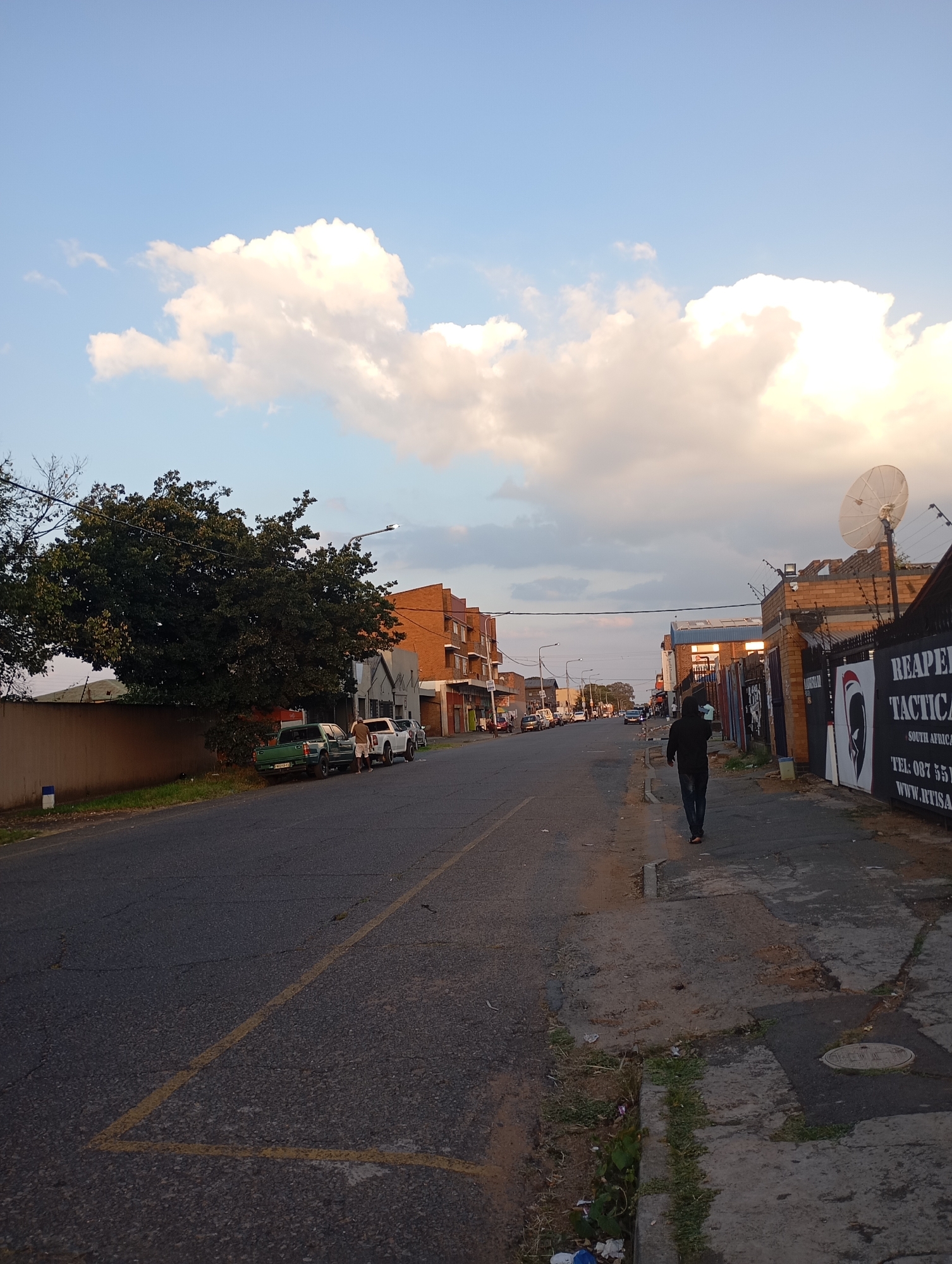 The image depicts a street in Johannesburg, South Africa. The street is paved and lined with buildings of varying heights and architectural styles, constructed primarily of brick. Several vehicles are parked on the left side of the street. On the right side of the street, a person wearing dark clothing is walking away from the camera. A satellite dish is mounted on the roof of a building on the right. The sky is partly cloudy with patches of blue visible.