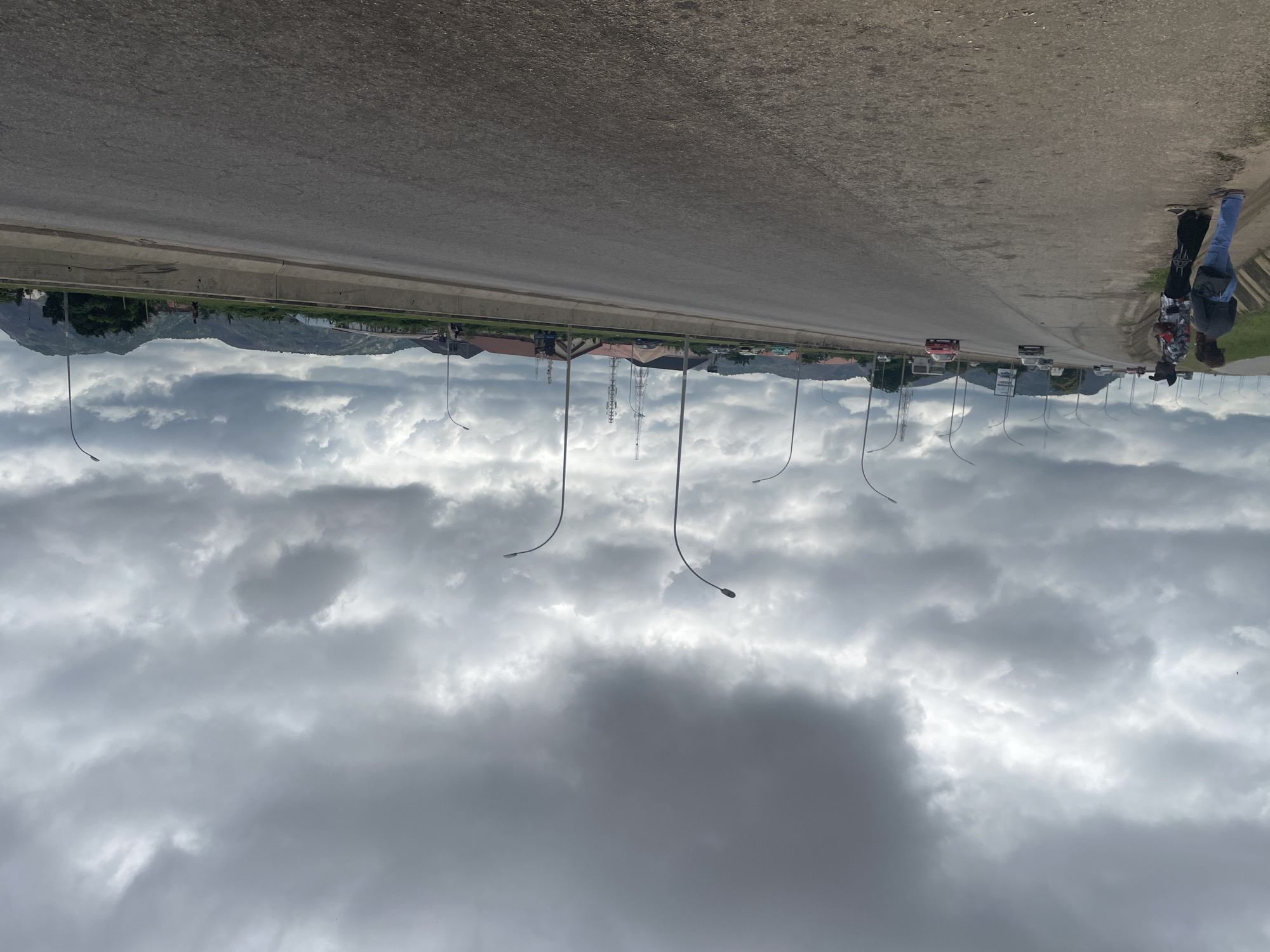 The image depicts a wide, gray asphalt highway under a cloudy, overcast sky. The highway is divided by a concrete barrier, and on the left side, a dirt shoulder is visible with two individuals standing. One is a woman in blue jeans and a dark top with a blue purse, and the other is a man wearing a patterned shirt, dark pants, and a black cap. Several vehicles are driving on the highway in both directions, including a red car. Streetlights with curved poles line the side of the road. In the distance, along the side of the highway, are buildings with red roofs, communication towers, and rolling hills covered in greenery. The overall scene suggests an urban or semi-urban environment in Kubwa, Nigeria, with the overcast sky indicating potential for rain.