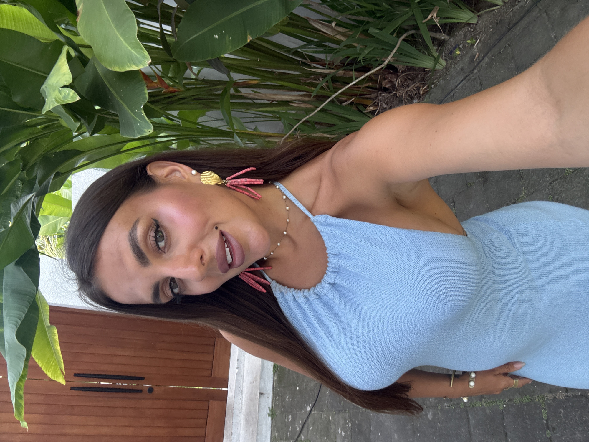 A woman is posing for a selfie outdoors in North Kuta, Indonesia. The woman has long brown hair, brown eyes, and wears a light blue dress, gold and red earrings, and bracelets. She is standing near a wooden door and lush greenery. The ground appears to be a paved walkway.