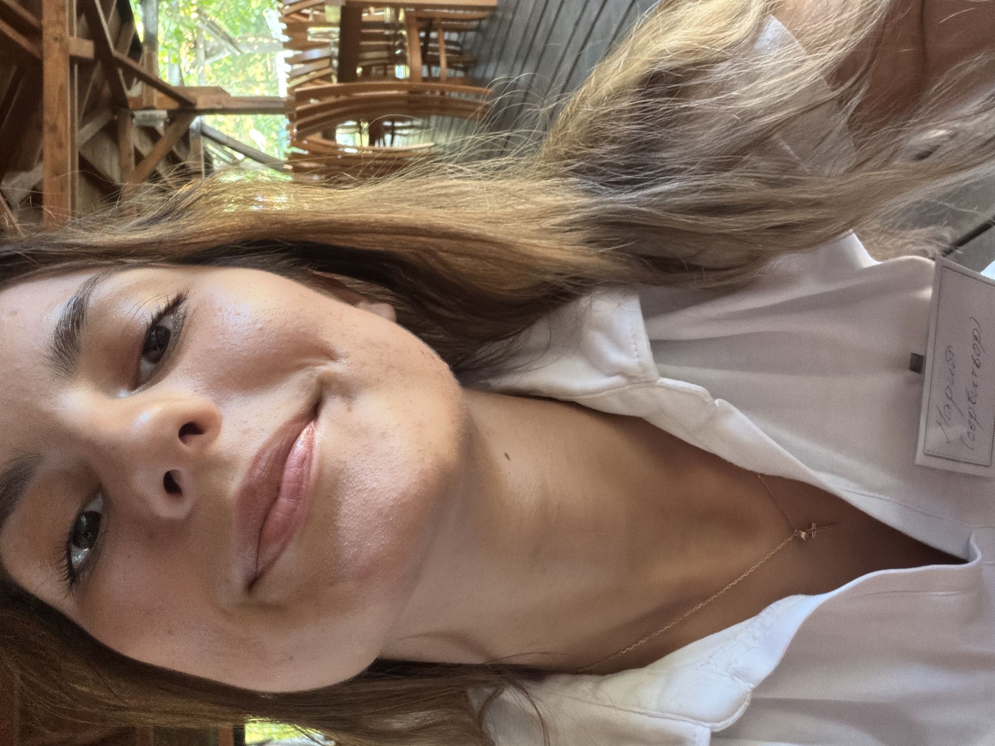 The image shows a close-up, selfie-style shot of a young woman with long, blonde hair and fair skin. She is wearing a white collared shirt and a delicate gold necklace with a cross pendant. A name tag is visible on her shirt, with handwritten text that appears to be in Cyrillic script. The background suggests an outdoor setting, possibly a restaurant or cafe with wooden furniture and visible foliage, consistent with the town of Tsarevo, Bulgaria, a coastal resort town known for its natural beauty and seaside dining establishments.