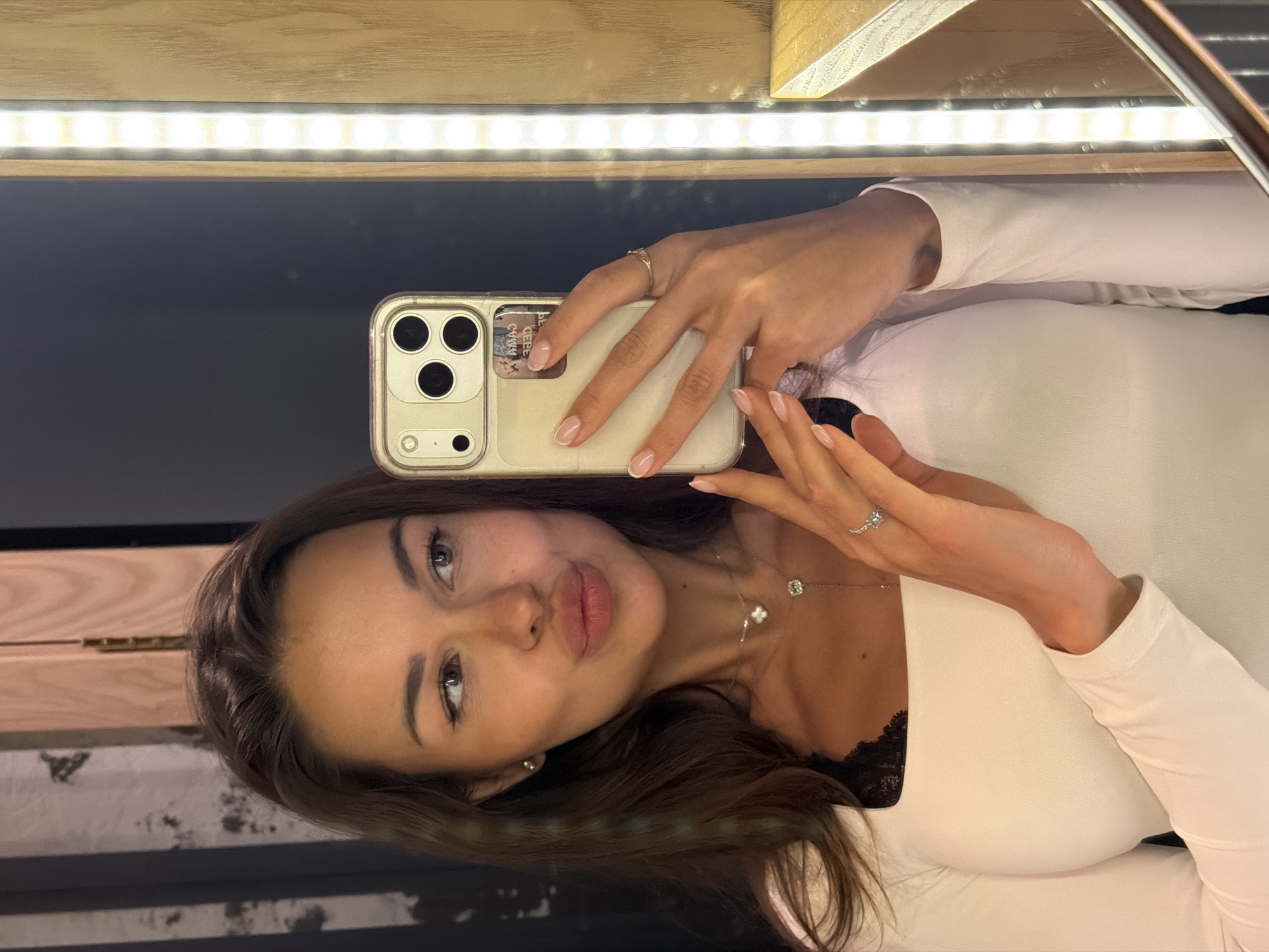 A young woman takes a mirror selfie in a dimly lit room. She has long, dark brown hair and is wearing a white long-sleeved top with a black lace trim. She is also wearing a delicate necklace with two charms and a ring on her left hand. The phone she is holding is a light-colored iPhone with a clear case. In the background, a vertical LED light strip is visible, illuminating part of the room and reflecting off the mirror. The environment suggests an indoor setting, possibly a dressing room or a bathroom, with wooden accents and dark walls. The image does not provide specific details about its location within Saint Petersburg, Russia.