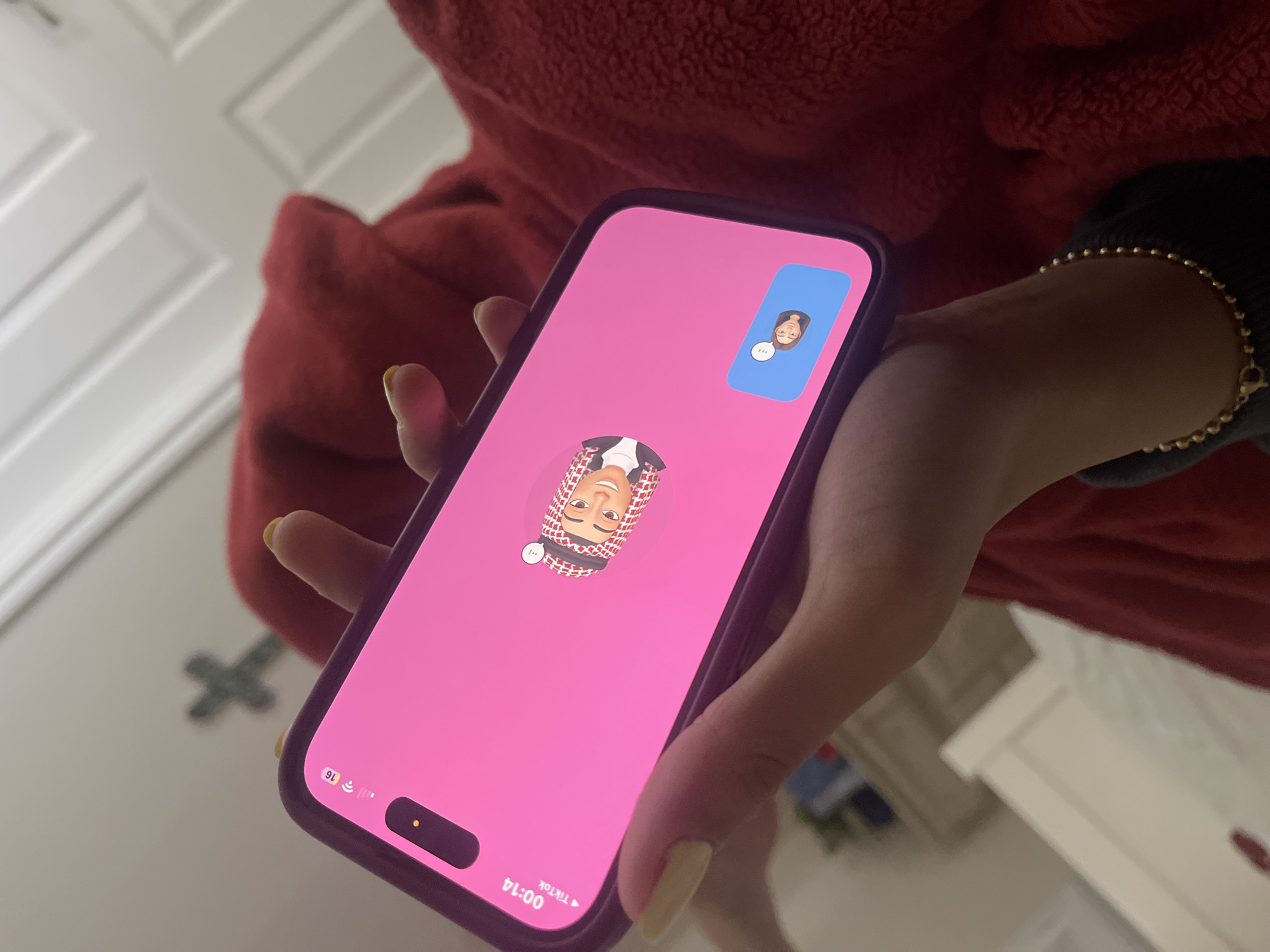A person is holding an iPhone showing a TikTok video call. The main video features an avatar of a person wearing a traditional Arab headdress, while the smaller inset video shows an avatar of a female-presenting person. The phone is held in front of a person wearing a red fleece hoodie. A gold beaded bracelet is visible on the person's wrist. The background includes a white door with recessed panels, a green four-pointed object resembling a stylized plus sign, and a white dresser or cabinet. The time on the phone is 00:14, and the cellular connection displays 5G. The location cannot be determined from this image.