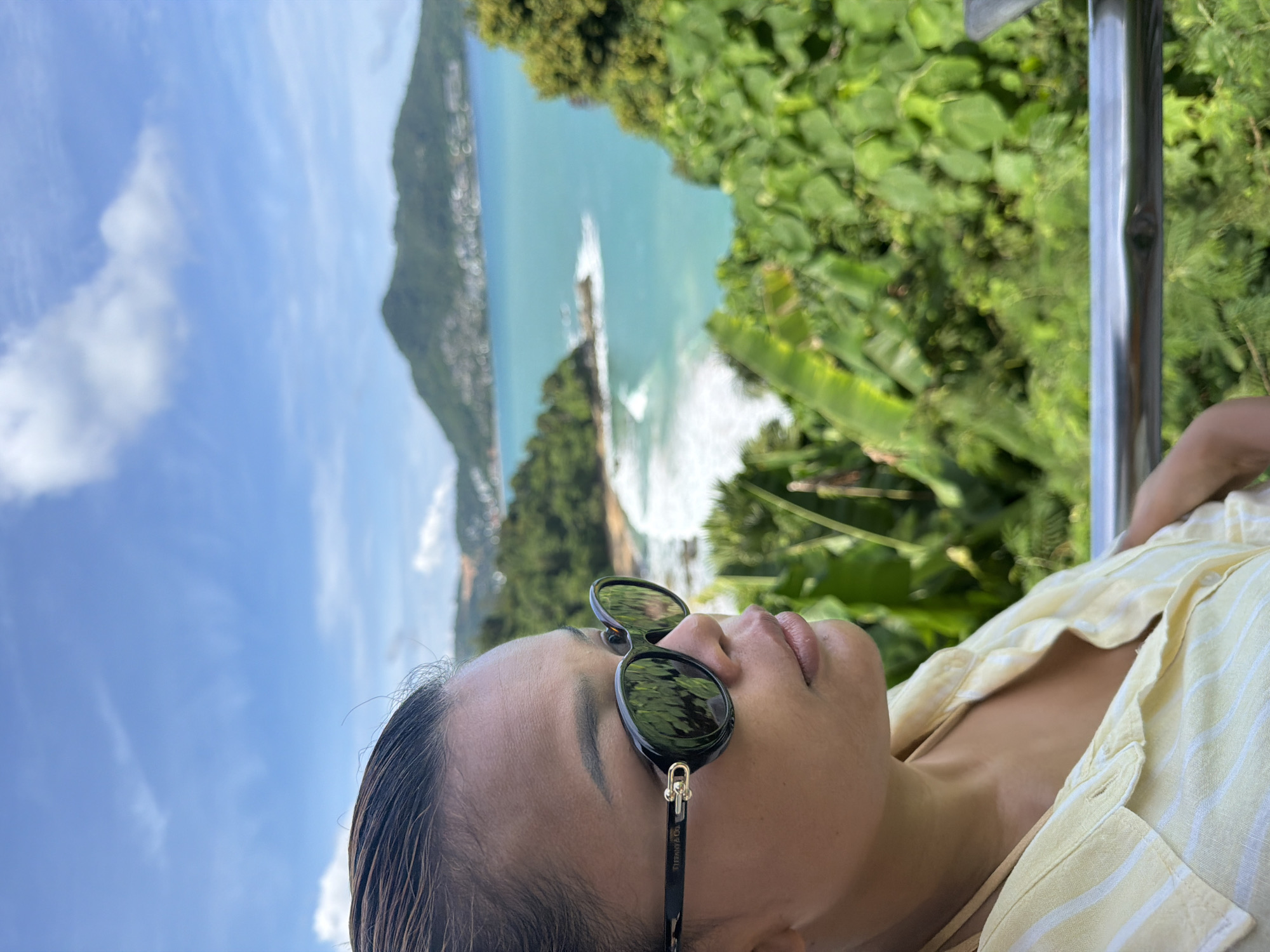 A medium shot captures a woman in profile, wearing sunglasses, against a backdrop of a bay with a curved coastline and lush green hills. The woman's hair is pulled back, and she is wearing a light-colored, striped shirt. The sunglasses are black with a tortoiseshell pattern on the arms and a gold accent. The bay features turquoise water with white waves crashing on a sandy beach. The hills are densely covered in vegetation, with some buildings visible on the slopes. The sky is blue with scattered white clouds. The scene is likely a scenic overlook in the Ban Bang Thao area of Thailand, known for its beaches and tropical landscape.