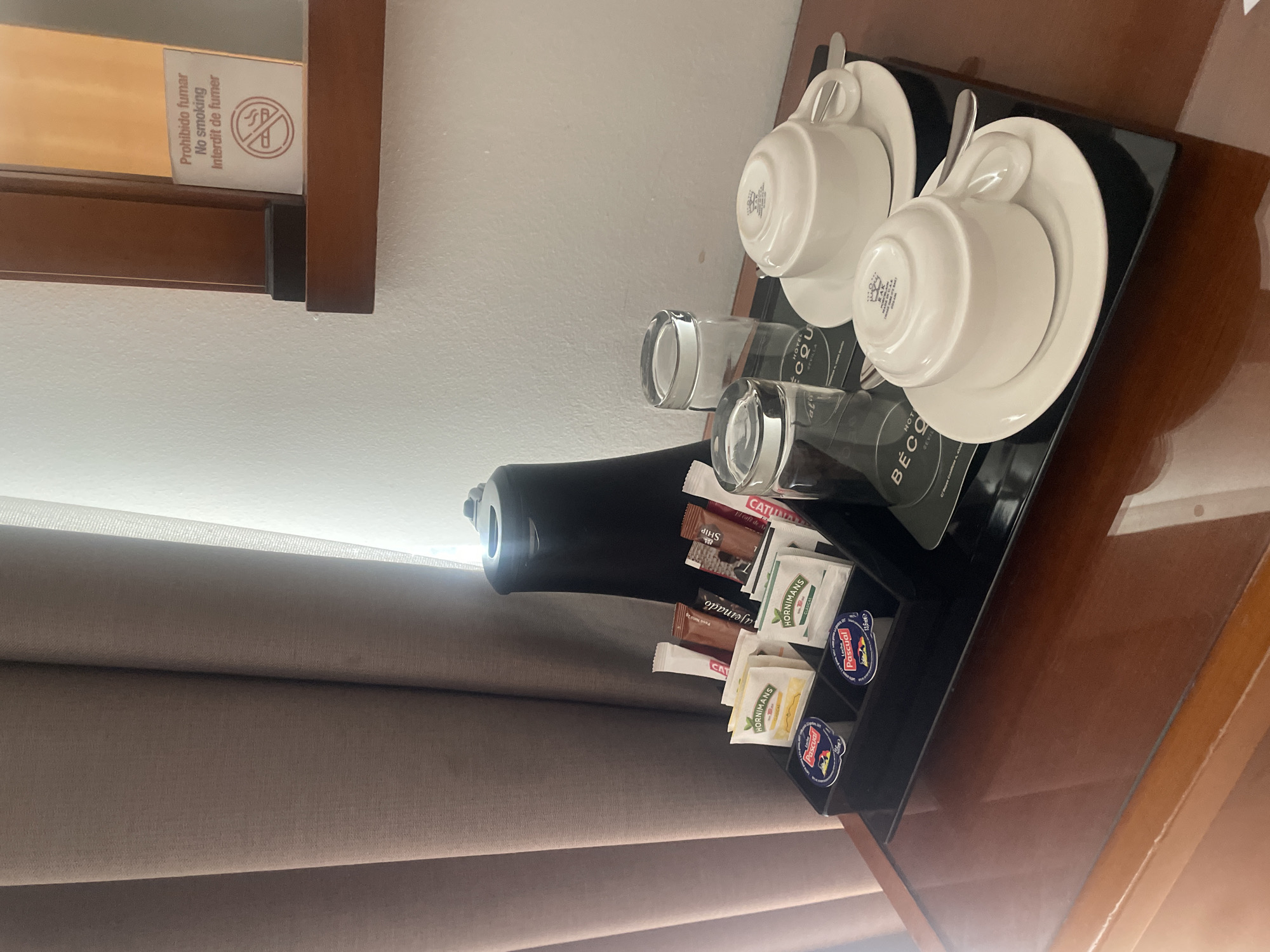 This is a high-angle, medium shot of a hotel room's beverage station. A dark brown wooden table holds a black tray with a collection of amenities. On the left, a black electric kettle is visible. To its right, within the tray, are various sachets of tea, coffee, and sugar, along with two small plastic tubs of "Leche Pascual." Two clear glass tumblers are also present. Further to the right, on saucers, are two white ceramic teacups, one upside down. A silver teaspoon rests on one of the saucers. A coaster with "HOTEL BECO SEVILLA" printed on it is under the tumblers and cups. In the background, a light-colored wall is partially obscured by a thick, brown curtain on the left and a thin, sheer white curtain on the right. On the wall, to the right, a wooden shelf holds a framed sign with Spanish, English, and French text stating "Prohibido fumar No smoking Interdit de fumer" along with a no-smoking symbol. The overall scene suggests a hotel room in Sevilla, Spain, prepared for guests to make hot beverages.