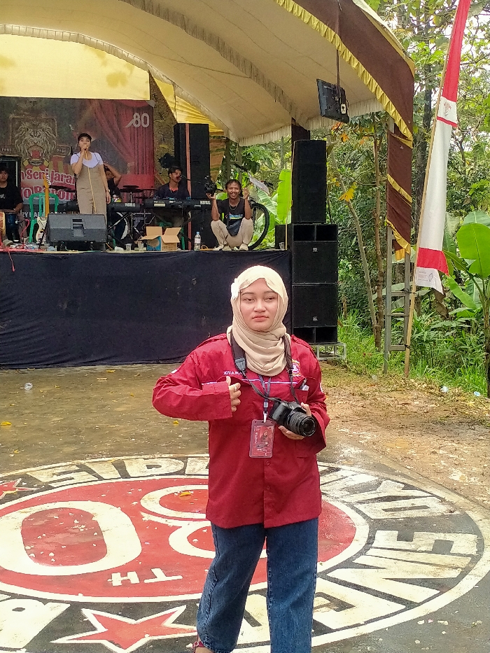A young adult female stands in the foreground, facing the camera with a neutral expression. She wears a beige hijab adorned with a small white flower on the left, a red long-sleeved collared shirt, and blue jeans. A black camera strap is around her neck, and she holds a black DSLR camera with a lens in her left hand. A red identification badge hangs from a lanyard on her right side, partially visible, with "JOVA PRATAMA" printed on her shirt. She has her left thumb hooked near her shirt pocket. She stands on a paved surface featuring a large circular red and white logo, which includes the number "80" and stylized text.
In the midground, a temporary outdoor stage is set up under a cream-colored tent canopy with a dark brown and gold valance. A large banner on the stage backdrop features a lion's head logo, text partially reading "PONPES SENJARA...", and the number "80." On the stage, a female performer in a light-colored dress stands at a microphone on the left, singing with her right hand raised. To her right, a male keyboardist is seated. Further right, a male videographer in a dark t-shirt and light pants squats, holding a video camera and smiling at the viewer. A bicycle is partially visible behind him. Black speaker stacks are positioned on both sides of the stage.
The background consists of dense green foliage, trees, and banana plants, indicating an outdoor, natural environment. To the right of the stage, a tall white and red Indonesian flag flies vertically on a pole. The scene takes place in the city of Tamansari, Indonesia.
