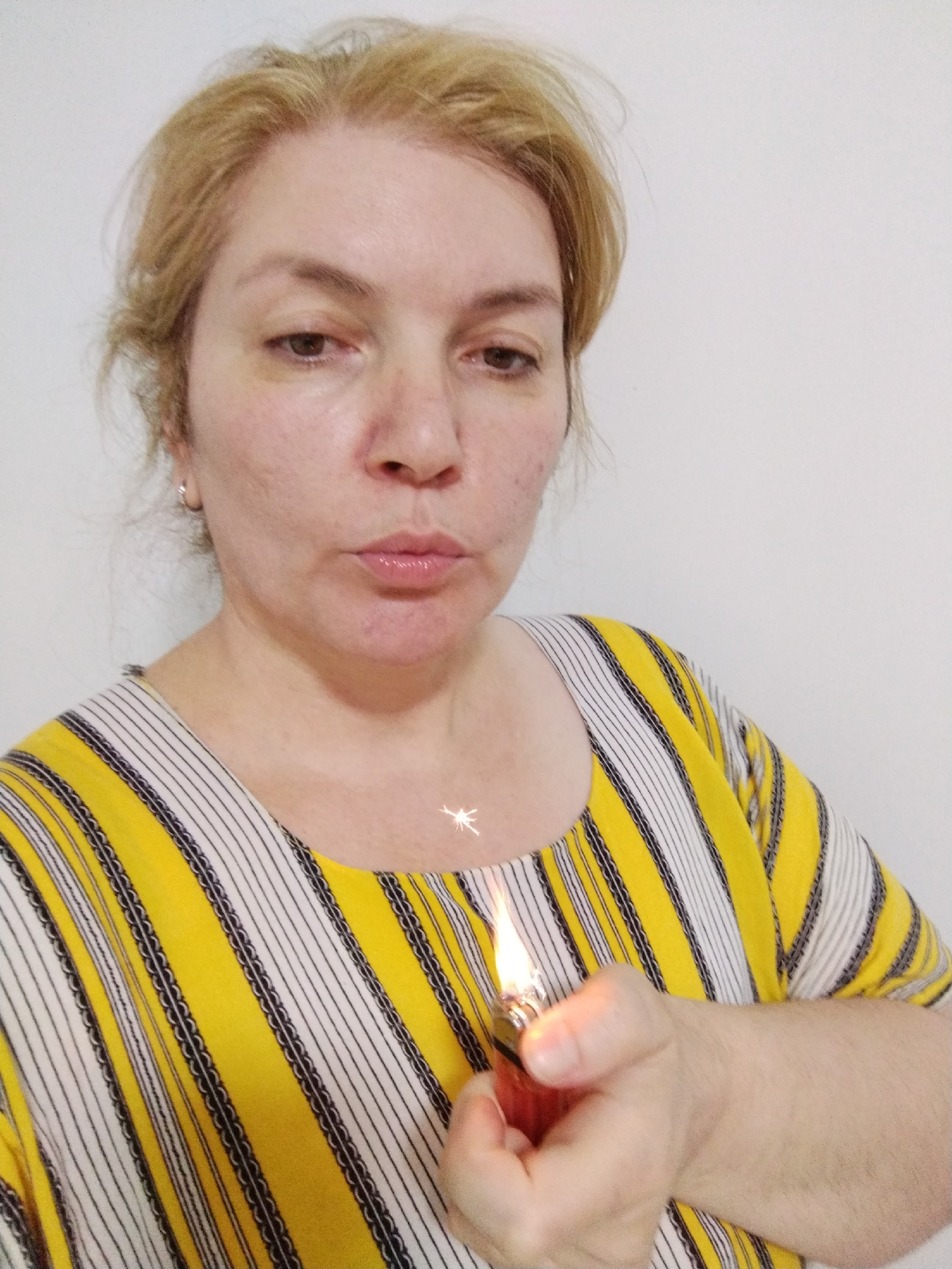 A woman is holding a lit lighter in her right hand, positioned near her chest. She has fair skin, blond hair, and is wearing a yellow and white striped shirt. The background is a plain white wall. The location is not evident in the image.