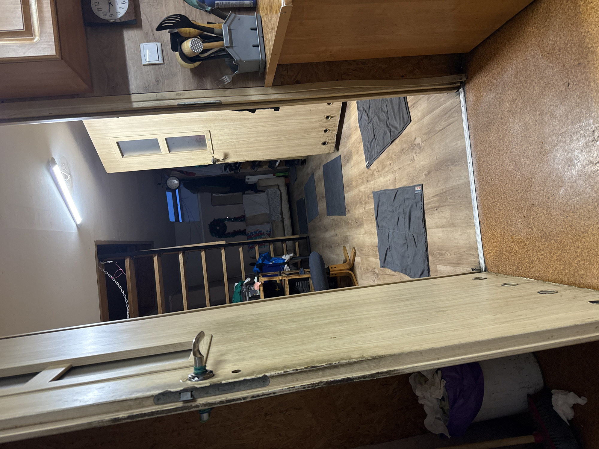 The image depicts an interior scene of what appears to be a residential dwelling. The foreground is dominated by a light-colored wooden door, partially open, revealing a hallway or living space beyond. The floor in the foreground is a textured, light brown material, possibly cork.
The hallway visible through the open door features a wooden floor with a visible grain. Several dark grey rectangular mats are laid out on the floor, spaced evenly. To the left, a set of wooden stairs leads upwards, with a metal chain attached near the top. A dark blue chair with a black cushion and wooden legs is positioned near the stairs. Shelving units are visible against the wall to the left of the stairs, holding various items.
In the background, through another doorway, a section of a kitchen area is visible on the right. This includes a wooden countertop, cabinetry, and various kitchen utensils in a holder. A clock with Roman numerals is mounted on the wall above the countertop. A light fixture is visible on the ceiling in the hallway.
A white trash can with a purple liner, containing white refuse, is situated in the lower left corner of the image, near a broom. The overall impression is a lived-in, somewhat cluttered interior space. There are no people visible in the image. The context provided about Wąwelnica, Poland, suggests this is an indoor scene within a residence in that location.