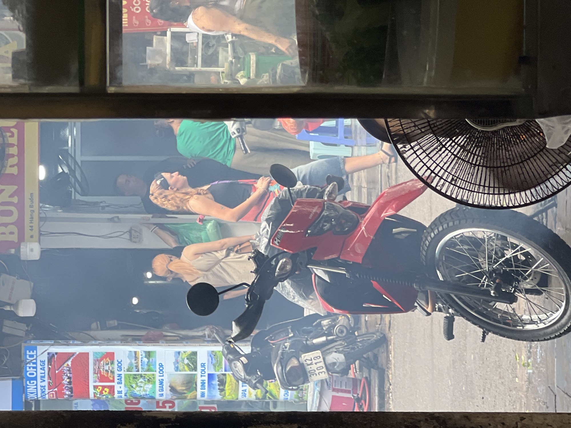 This is a scene from a street in Hanoi, Vietnam. In the foreground, a red motorcycle is parked, its front wheel and handlebars visible. To its left, another motorcycle is parked, showing a license plate that reads "30-K2 3018". Behind the motorcycles, a group of people are visible in the hazy street. On the left, a poster displays various tourist destinations. Further back, a woman with blonde hair pulled into a ponytail is wearing a light-colored tank top and jeans, talking to a woman with curly blonde hair wearing a black tank top. Another person in a green shirt is standing behind them. The street appears to be bustling with activity, and the overall atmosphere is hazy, possibly due to smoke or dust. A part of a fan is visible in the lower right corner of the image.