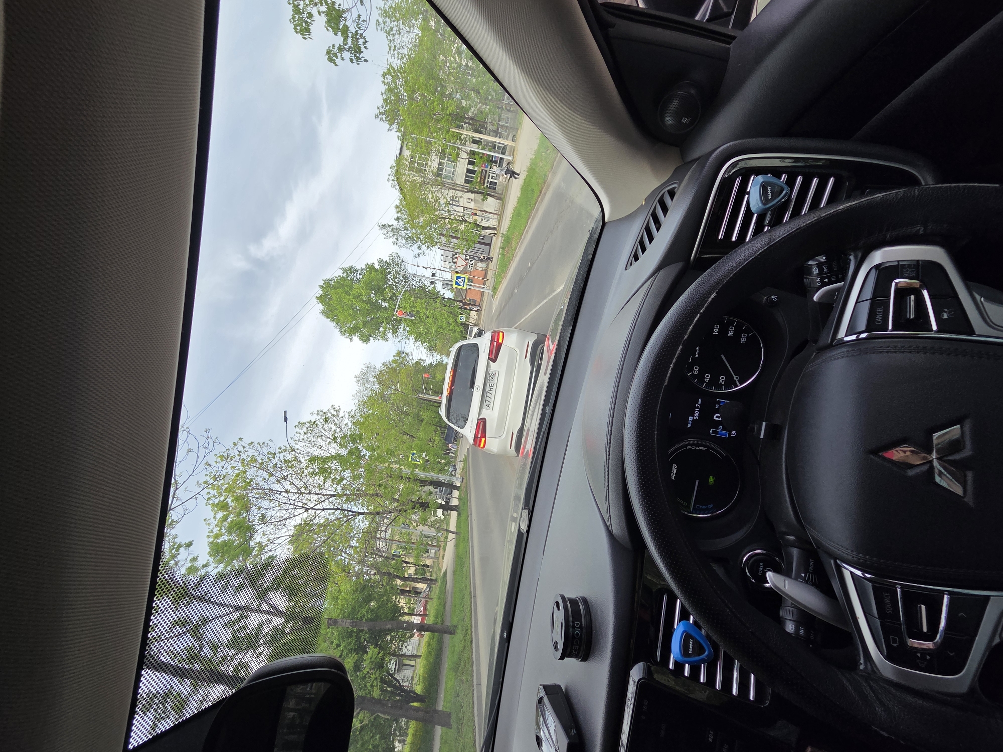 Captured from within a vehicle, the image presents a view of a city street. A white car is directly ahead, partially obstructing the view. The license plate "A777HE 125" is visible. A traffic light is discernible in the distance, with trees lining the street on either side. A building stands across the street, with a pedestrian walking in front of it. The interior of the car, including the steering wheel with the Mitsubishi logo, is in focus. Based on the license plate and general appearance, this image was taken in Birobidzhan, Russia.