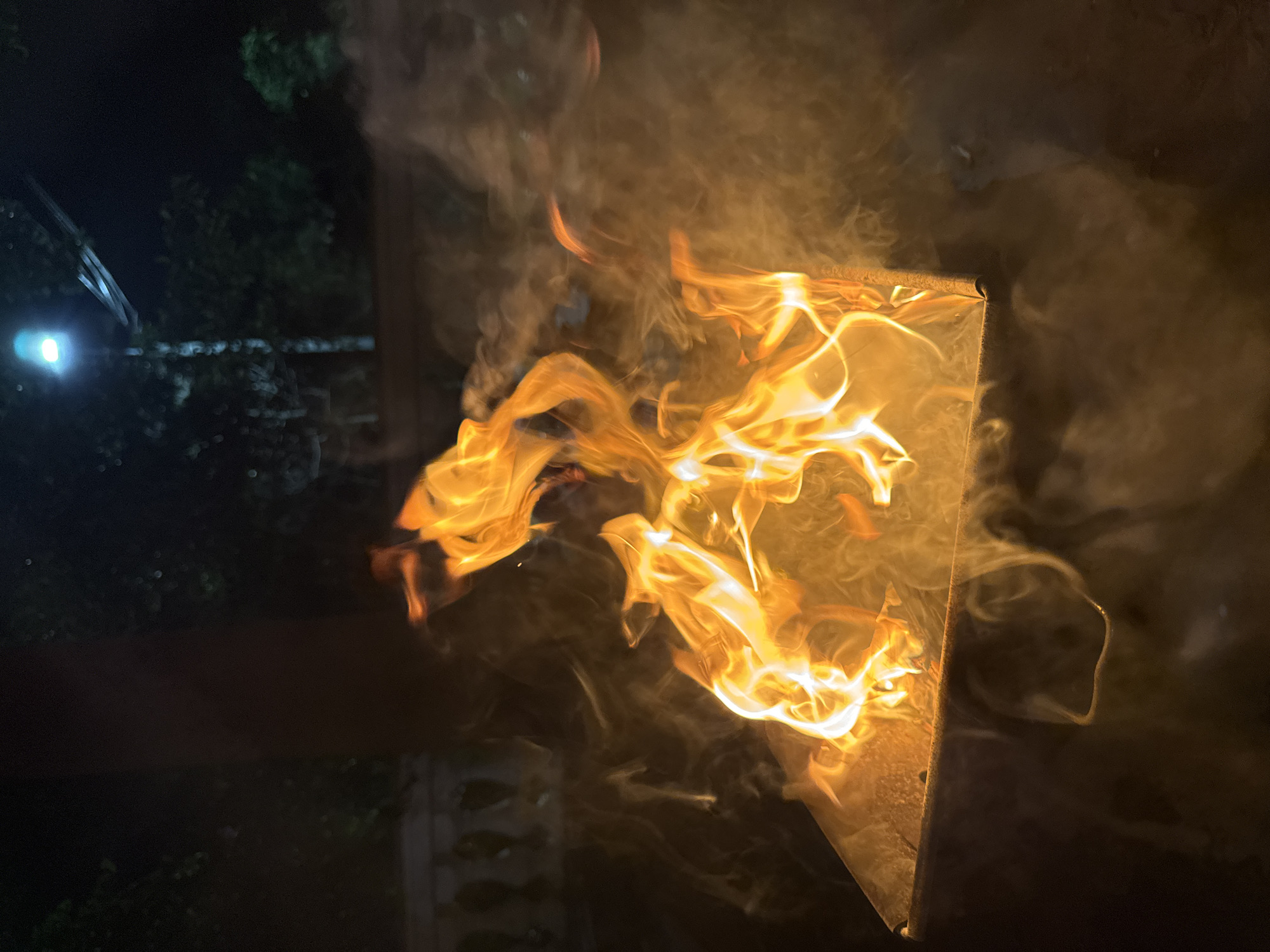 A nighttime outdoor scene in Mũi Né, Vietnam, captures a fire burning in a metal brazier. The flames are bright orange and yellow, with visible smoke tendrils swirling around them. In the background, a lamppost illuminates the dark sky, casting a soft glow on the surrounding trees and foliage. A white balustrade is partially visible to the left, suggesting a patio or garden setting. The overall atmosphere is warm and inviting, despite the darkness of the night.