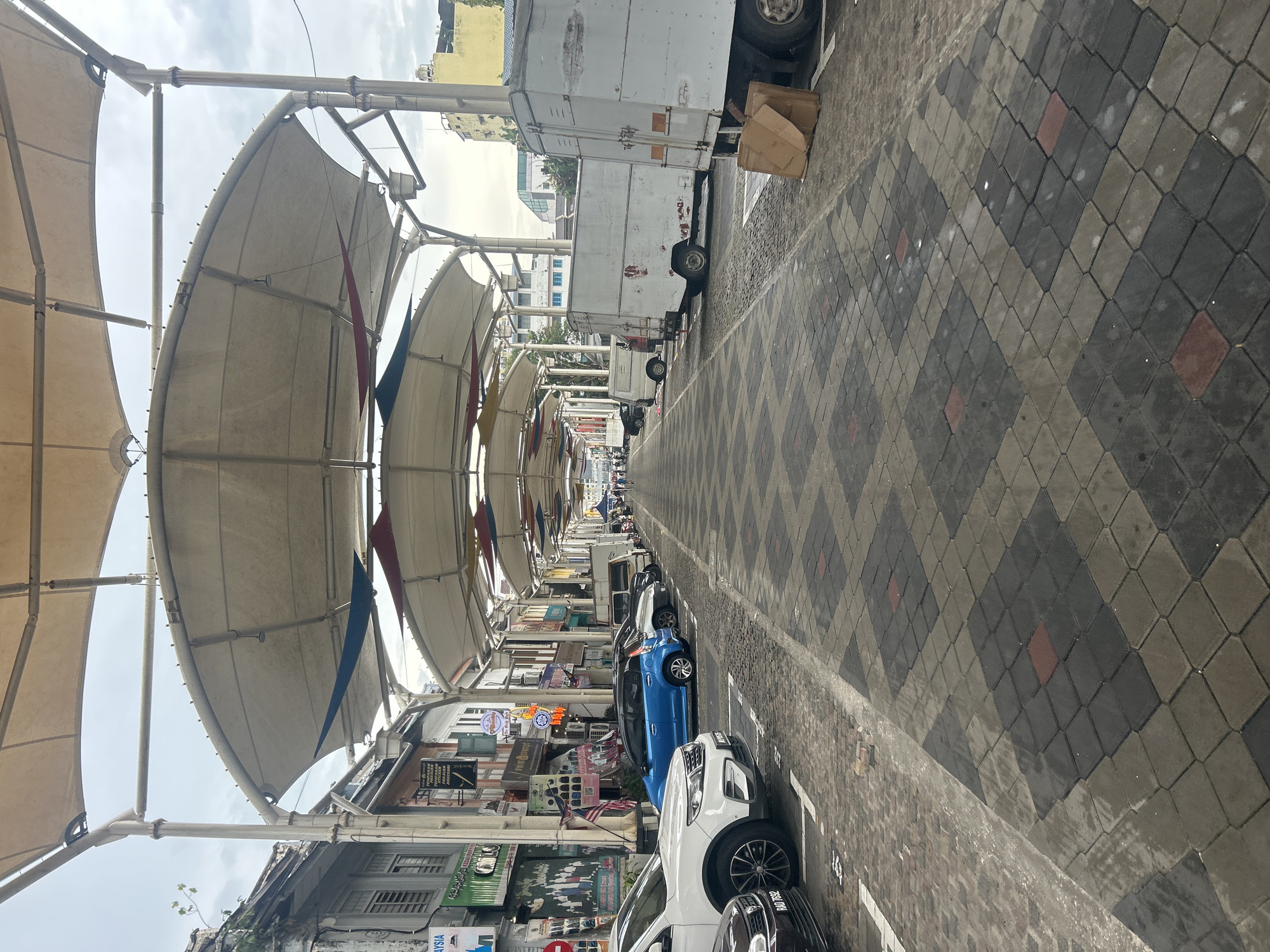 The image depicts a long, covered street or marketplace in Ipoh, Malaysia, likely a heritage area given the architecture of the buildings lining the sides. The street is covered by a series of large, white fabric canopies supported by metal poles, creating a sheltered walkway. The ground is paved with patterned interlocking stones in shades of grey and dark grey, with some red accents, and appears to be a parking area as several cars are parked along the left side of the street. On the right side, there are several parked delivery trucks and vans, some showing signs of wear and tear, with cardboard boxes piled near one of them. The buildings on the left showcase traditional shop fronts with signage in Malay and possibly Chinese. The overall atmosphere is that of a bustling commercial area, perhaps a market or a street with shops and eateries, adapted with coverings and parking facilities. A few individuals can be seen walking in the distance on the right side of the street. The sky is overcast, suggesting a cloudy day.