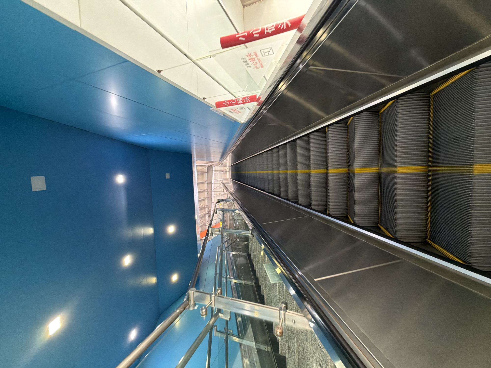 The image shows a view from the bottom of an escalator looking upwards. The escalator has dark grey steps with yellow safety lines. To the left of the escalator are stairs with a modern railing system made of glass and metal. The ceiling is a bright, vibrant blue, with several rectangular lights integrated into it. The walls are also blue, with some white panels. There are red signs with Chinese text and an English warning that reads "Mind your head" hanging on the right side of the escalator. The scene appears to be a public transportation hub or a modern building in Tiaodeng, China. There are no people clearly visible on the escalator or stairs, but the setting suggests a place with regular foot traffic. The lighting is bright and even, creating a clean and contemporary atmosphere.
