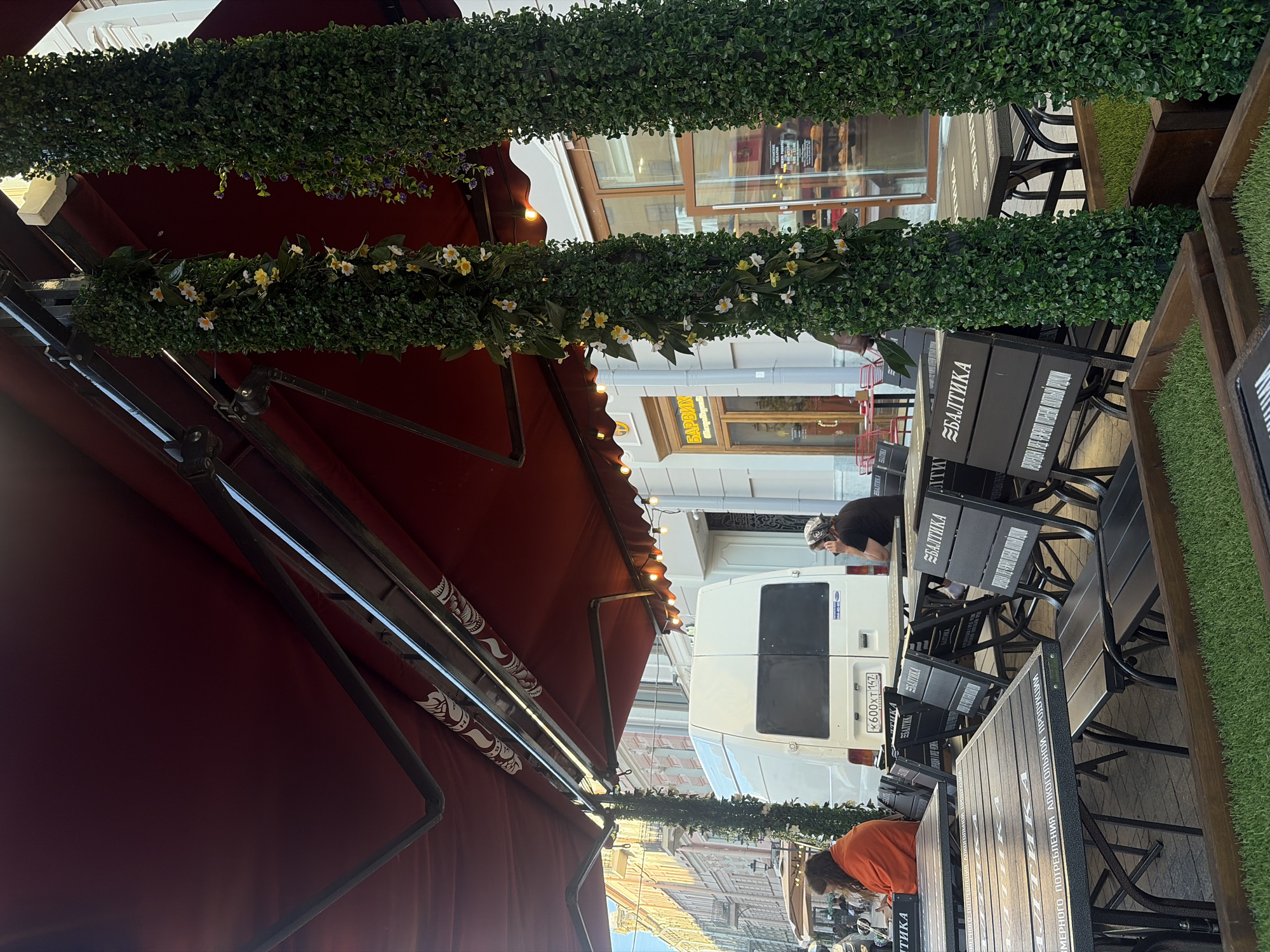 An outdoor seating area of a restaurant or cafe is depicted, likely in an urban setting. The scene is shaded by a large, dark red awning supported by metal beams. Two tall, cylindrical structures covered in artificial greenery and small white and yellow flowers are prominent features, positioned at the edge of the seating area. In the background, a white van is parked, displaying a license plate that reads "K600XT 147". Adjacent to the van is a building with large windows and signage that includes the word "БАРИХ". Several tables and chairs, some with the "БАЛТИКА" logo visible on the backs, are arranged on the pavement. In the foreground, more tables and chairs are set up, with planters containing artificial grass. Two individuals are seated at tables: one person in an orange garment is at a table on the left, facing away from the camera, and another person wearing a dark shirt and headscarf is seated at a table on the right, facing the side. The lighting suggests it is daytime, with sunlight illuminating parts of the scene. The overall impression is of a pleasant, possibly European, outdoor dining environment.