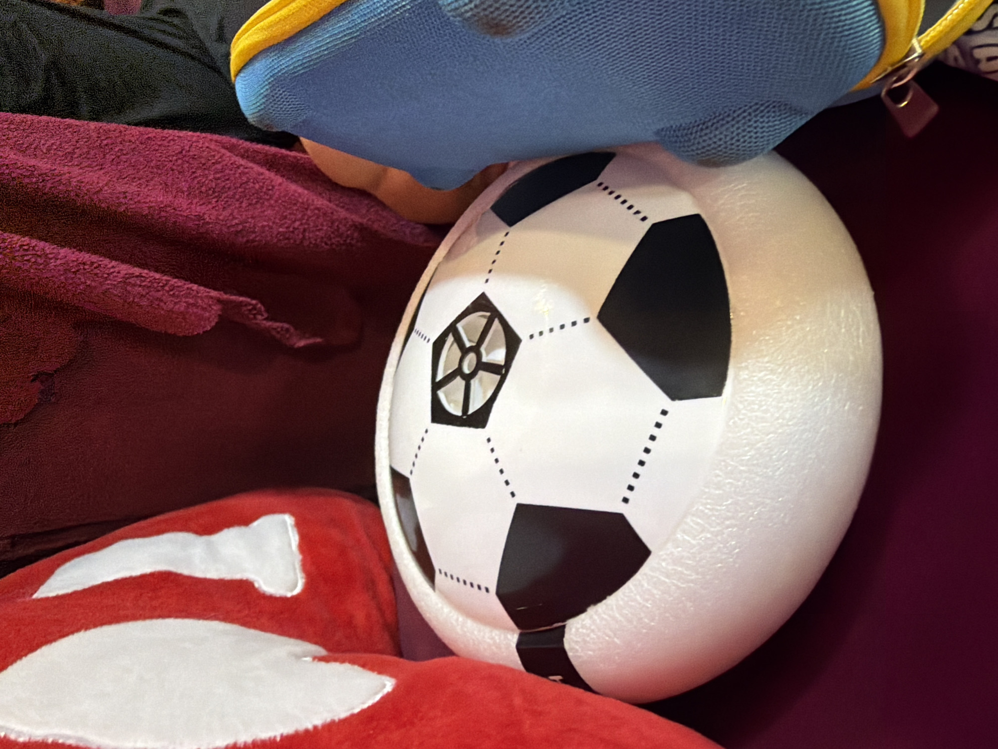 A close-up, slightly angled shot captures a white and black patterned soccer ball nestled among various soft fabrics. The soccer ball is prominently displayed, featuring the classic hexagonal and pentagonal patterns, with white main panels and black accents. One central black pentagonal section has an intricate, star-like design in white. The surface of the white panels appears textured, possibly indicative of foam or a similar material, suggesting it might be a decorative item or a lightweight toy.
Surrounding the ball are multiple textiles. In the foreground and bottom left, a vibrant red, plush fabric, likely a pillow or blanket, is visible with distinct white, abstract shapes on its surface. Behind the soccer ball, a rich, dark red or maroon textured fabric, also appearing soft and plush, extends across the mid-ground. Further up and to the left, a piece of black fabric is partially visible. At the top, a blue item with a mesh-like texture, possibly a bag or a backpack, rests above the soccer ball. It features a bright yellow zipper and a small, metallic zipper pull or tag.
The scene appears to be an indoor setting, possibly a bed or a cozy corner, with the items arranged casually. The lighting is soft and even, without harsh shadows, and no specific time of day or weather conditions can be determined. There is no visible text on any of the objects.
