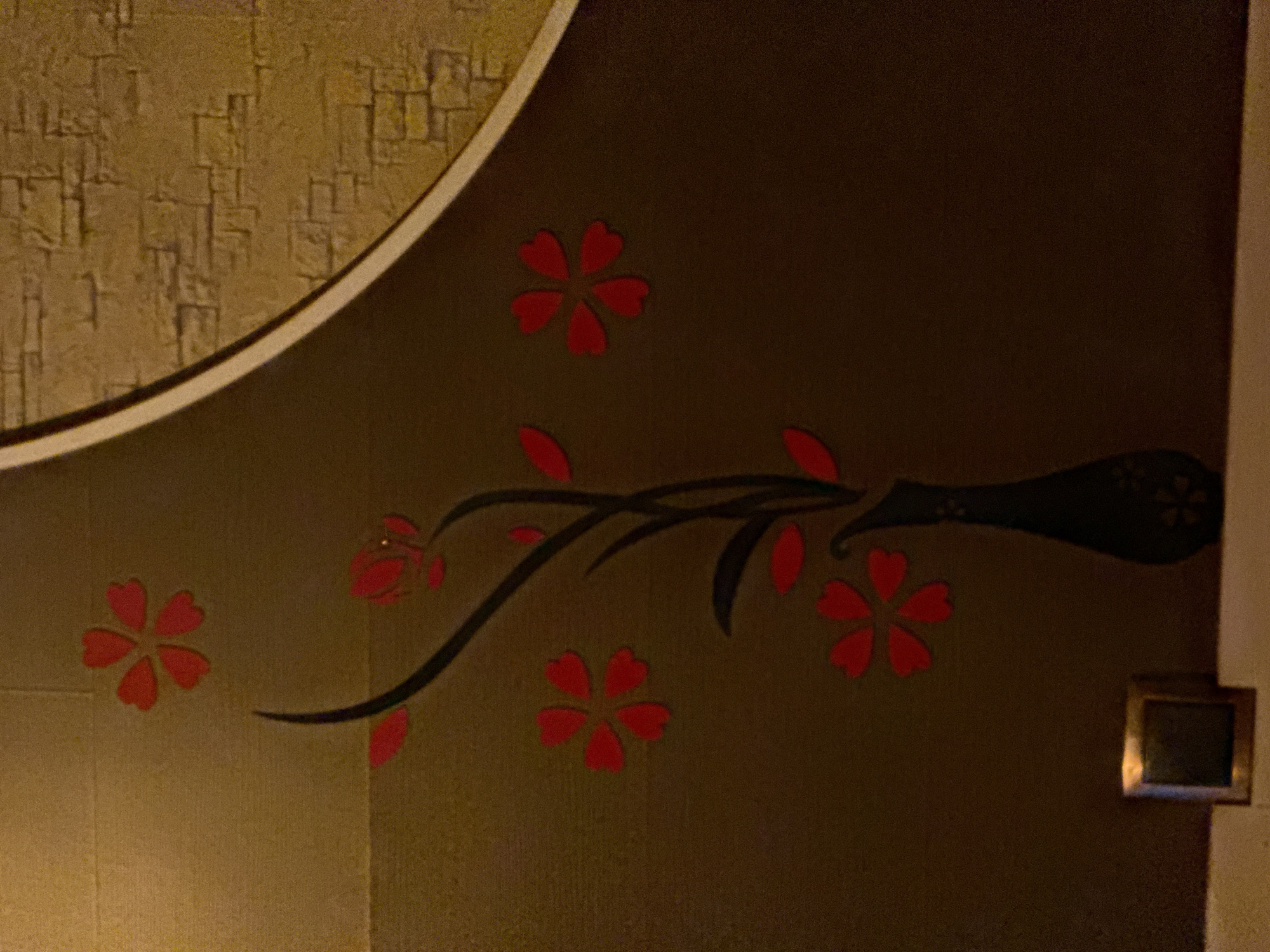 A decorative wall feature in the city of Galinė, Lithuania, depicts a stylized black branch with red blossoms. The branch emerges from a dark vase and extends upwards, with multiple red, five-petaled flowers scattered around it. To the right of the floral motif, a section of the wall features a textured pattern resembling light brown bricks, bordered by a curved, cream-colored accent. In the lower left corner, a small, metallic square object rests on a light-colored surface.
