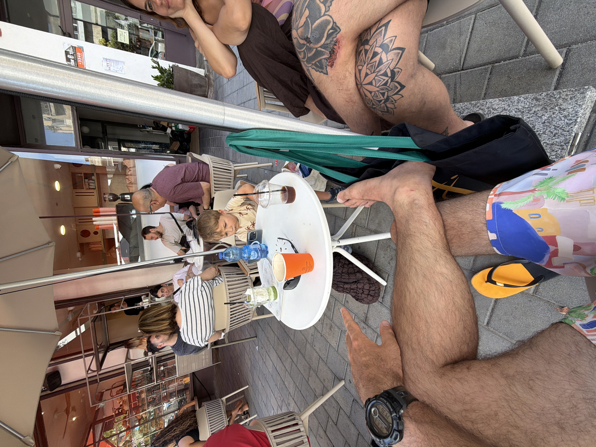 The image captures an outdoor cafe scene with people relaxing at tables, likely in Tel-Aviv, Israel given the context. The time of day appears to be daytime, with ample natural light suggesting a sunny day.
In the foreground, a person's bare legs are visible, one with intricate tattoos (a rose and a mandala pattern). This person is wearing colorful shorts and an orange flip-flop is on the ground nearby. Another person's arm with a watch on the wrist is also in the foreground, pointing towards a white round table.
At the white table, several items are placed: a plastic cup with a straw containing a dark beverage (possibly coffee or a smoothie), a blue water bottle, an orange paper cup, and sunglasses. A young boy with blonde hair is seated at this table, looking at something off-camera.
Further back, more individuals are seen seated at other cafe tables. One man with glasses and a purple shirt is leaning over the table. Another woman wears a black and white striped shirt. Reflections of buildings and the street are visible in the windows of the establishments across the cafe. The ground is paved with gray tiles.
Notable details include the various tattoos, the casual attire of the people, and the typical cafe setting with outdoor seating. The overall mood appears relaxed and social. The visible text on a building in the background includes "3340" and some Hebrew characters, supporting the Tel-Aviv location context.